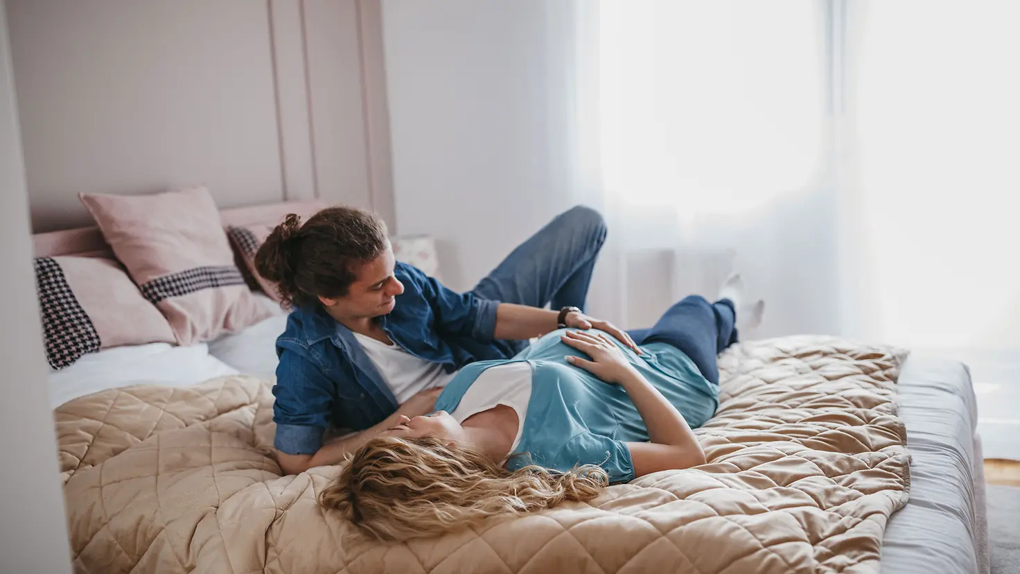 Junges Paar liegt auf einem Bett und streicheln den Babybauch der etwa 23. Schwangerschaftswoche
