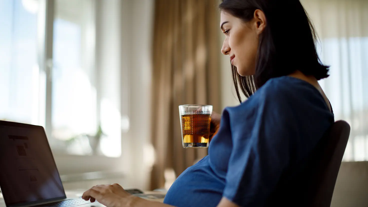 Schwangere Frau in der etwa Schwangere Frau in der etwa 24. Schwangerschaftswoche hält Gabel mit Käse in der Handsitzt am Computer und hält ein Teeglas in der Hand