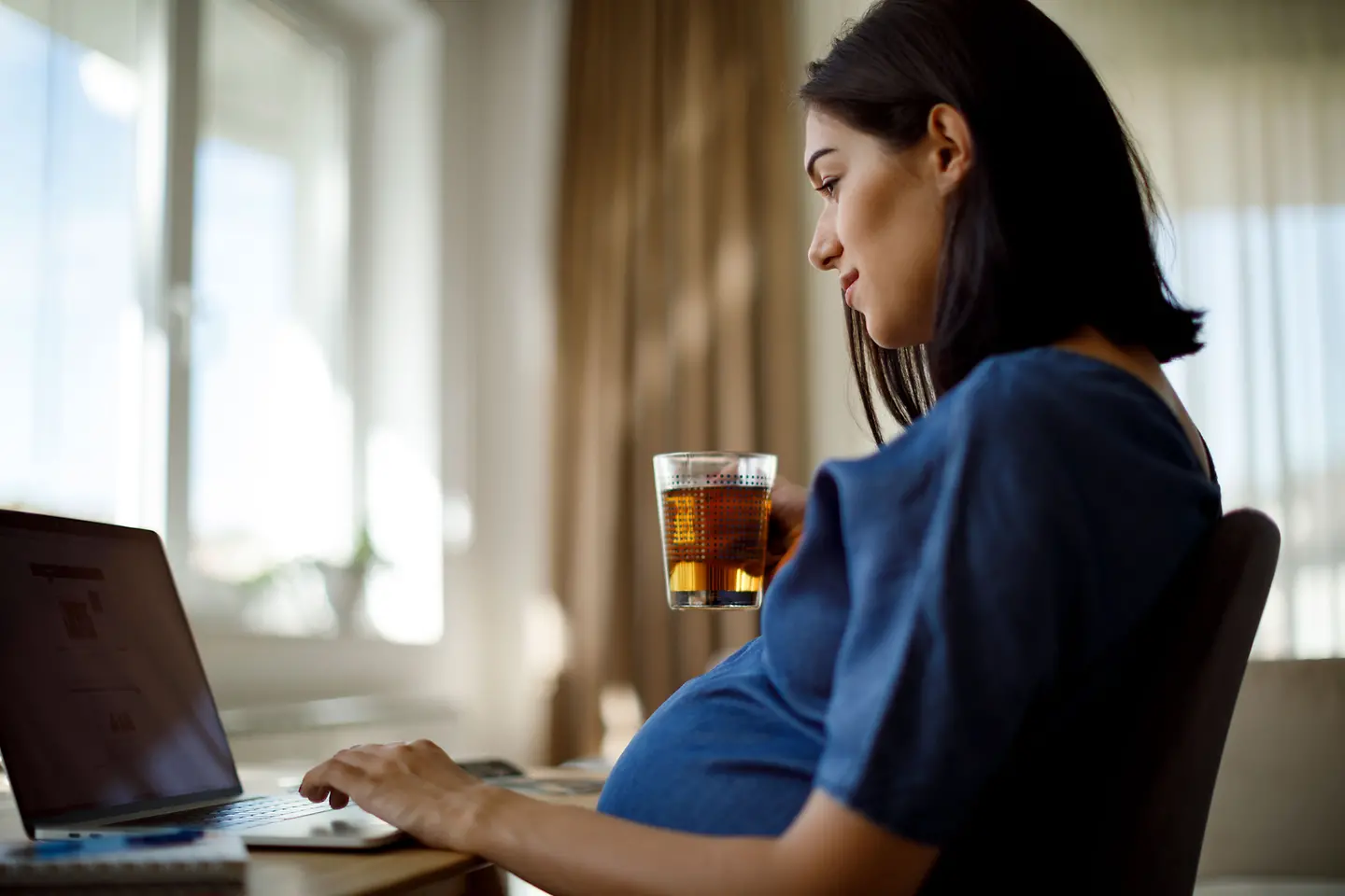 Schwangere Frau in der etwa Schwangere Frau in der etwa 24. Schwangerschaftswoche hält Gabel mit Käse in der Handsitzt am Computer und hält ein Teeglas in der Hand