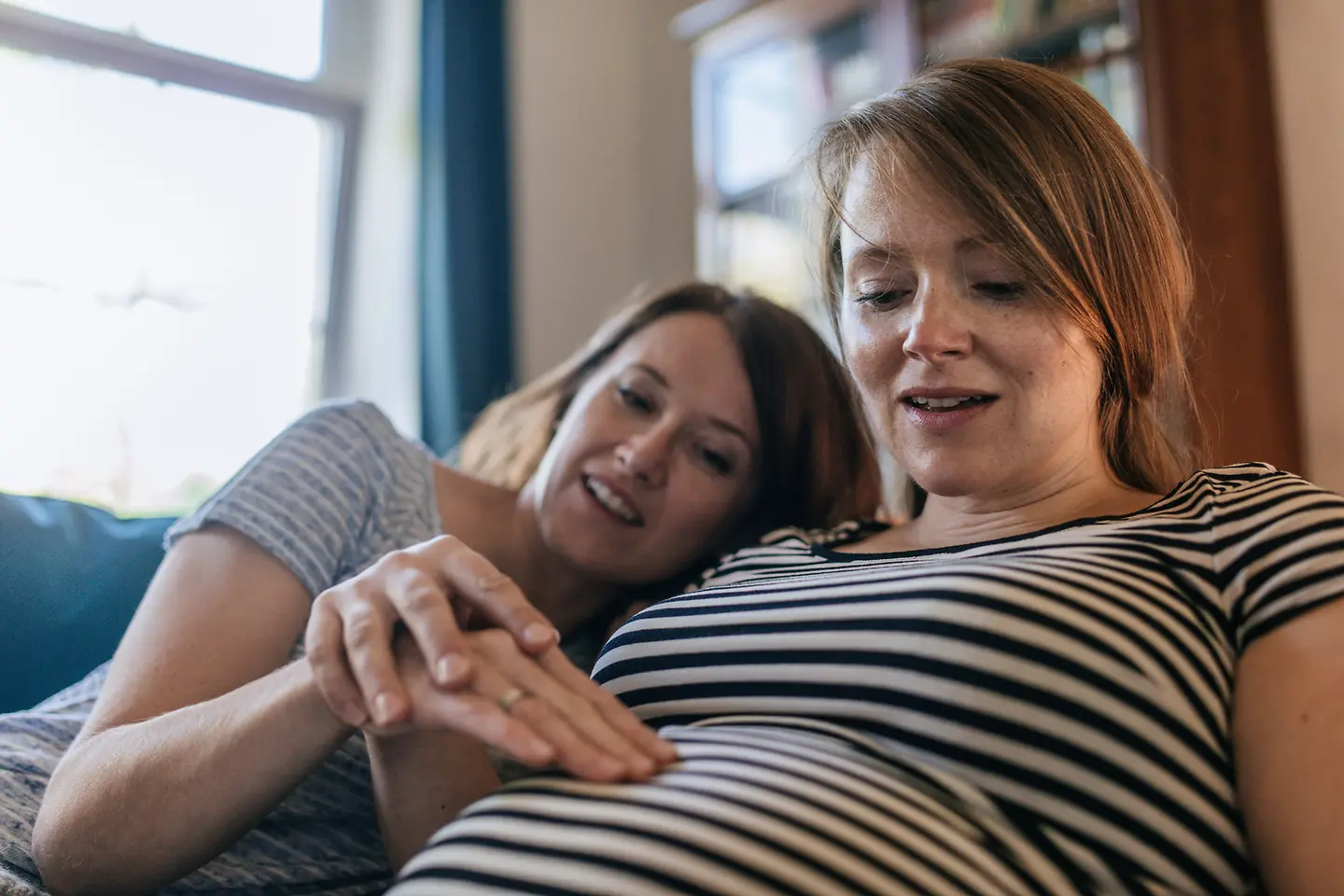 Eine Frau in der etwa 25. Schwangerschaftswoche führt die Hand einer anderen Frau, die ihren Babybauch abtastet