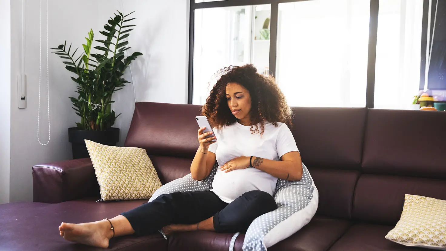 Schwangere Frau in der etwa 25. Schwangerschaftswoche sitzt auf der Couch und schaut ihr Mobiltelefon
