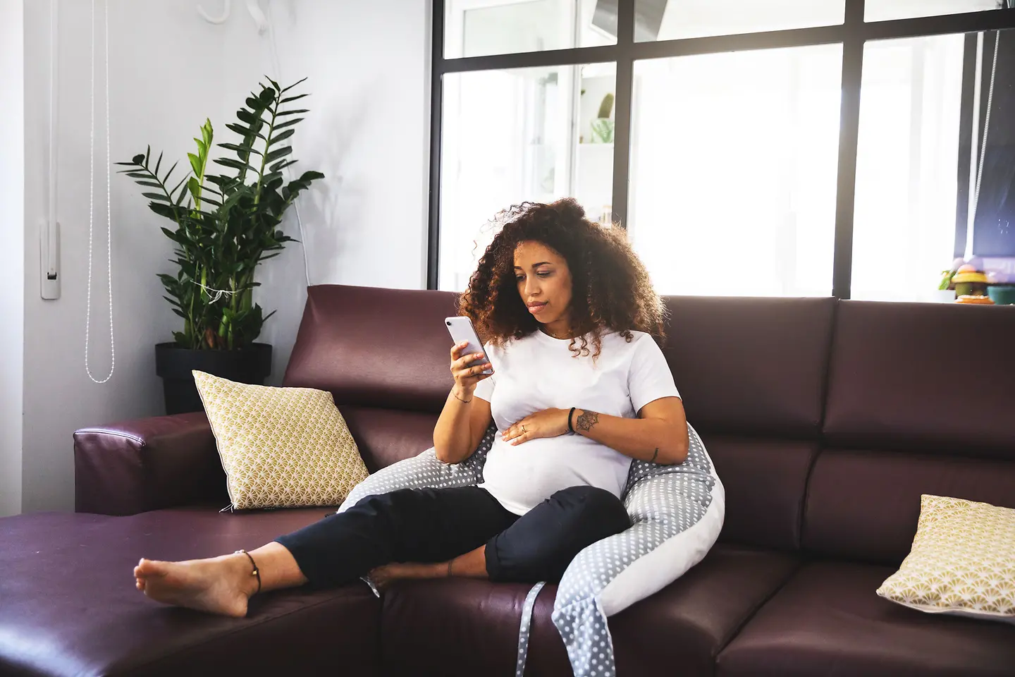 Schwangere Frau in der etwa 25. Schwangerschaftswoche sitzt auf der Couch und schaut ihr Mobiltelefon