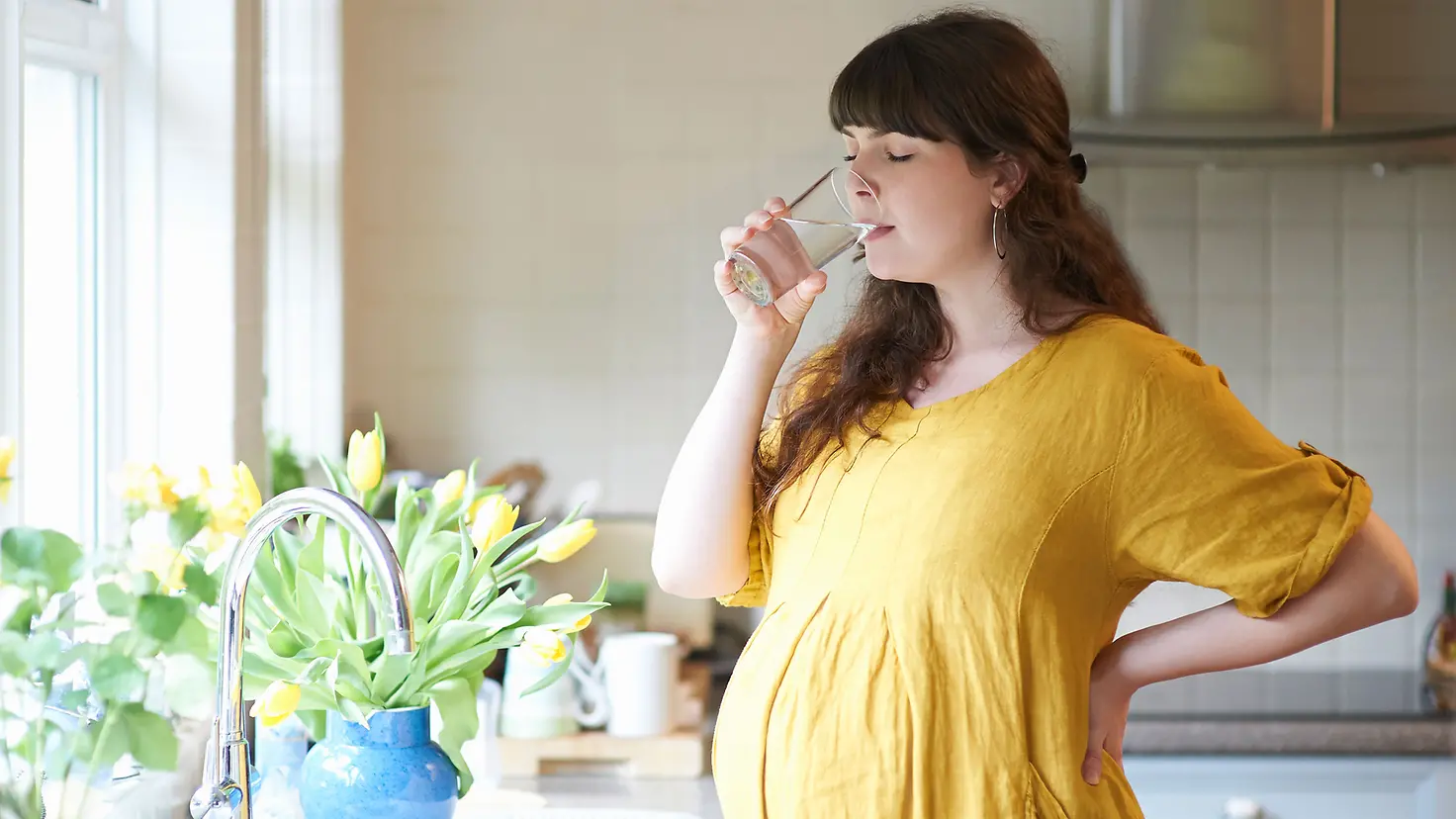 Schwangere Frau um die 30. Schwangerschaftswoche trinkt ein Glas Wasser an Fenster in der Küche