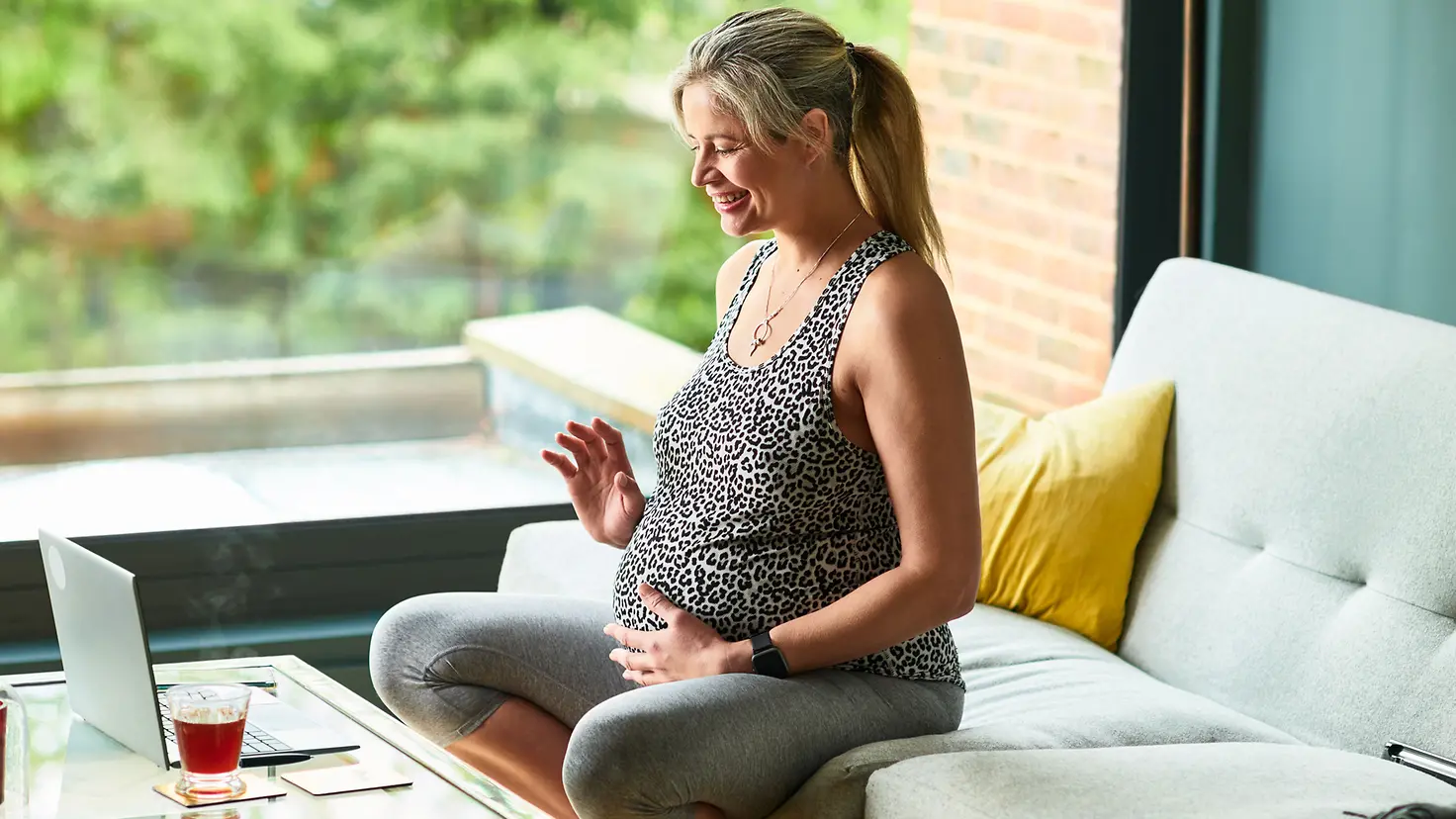 Schwangere Frau in der etwa 26. Schwangerschaftswoche sitzt auf dem Sofa mit der Hand auf dem Bauch, spricht und winkt in die Webcam eines Laptops