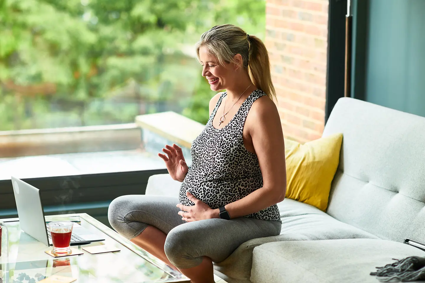Schwangere Frau in der etwa 26. Schwangerschaftswoche sitzt auf dem Sofa mit der Hand auf dem Bauch, spricht und winkt in die Webcam eines Laptops