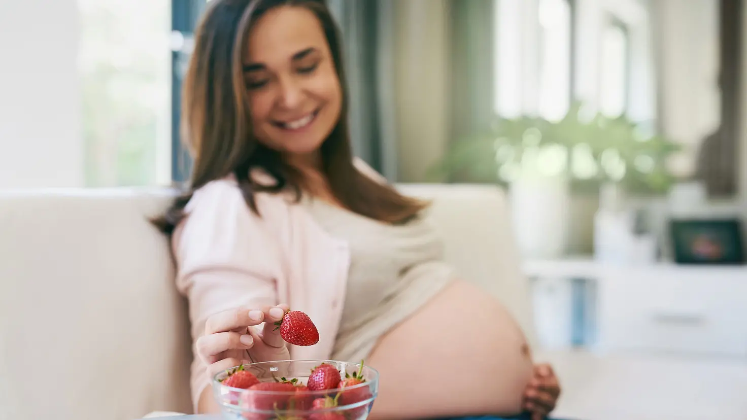 Schwangere Frau in der etwa 27. Schwangerschaftswoche isst Erdbeeren und hält sich den nackten Babybauch