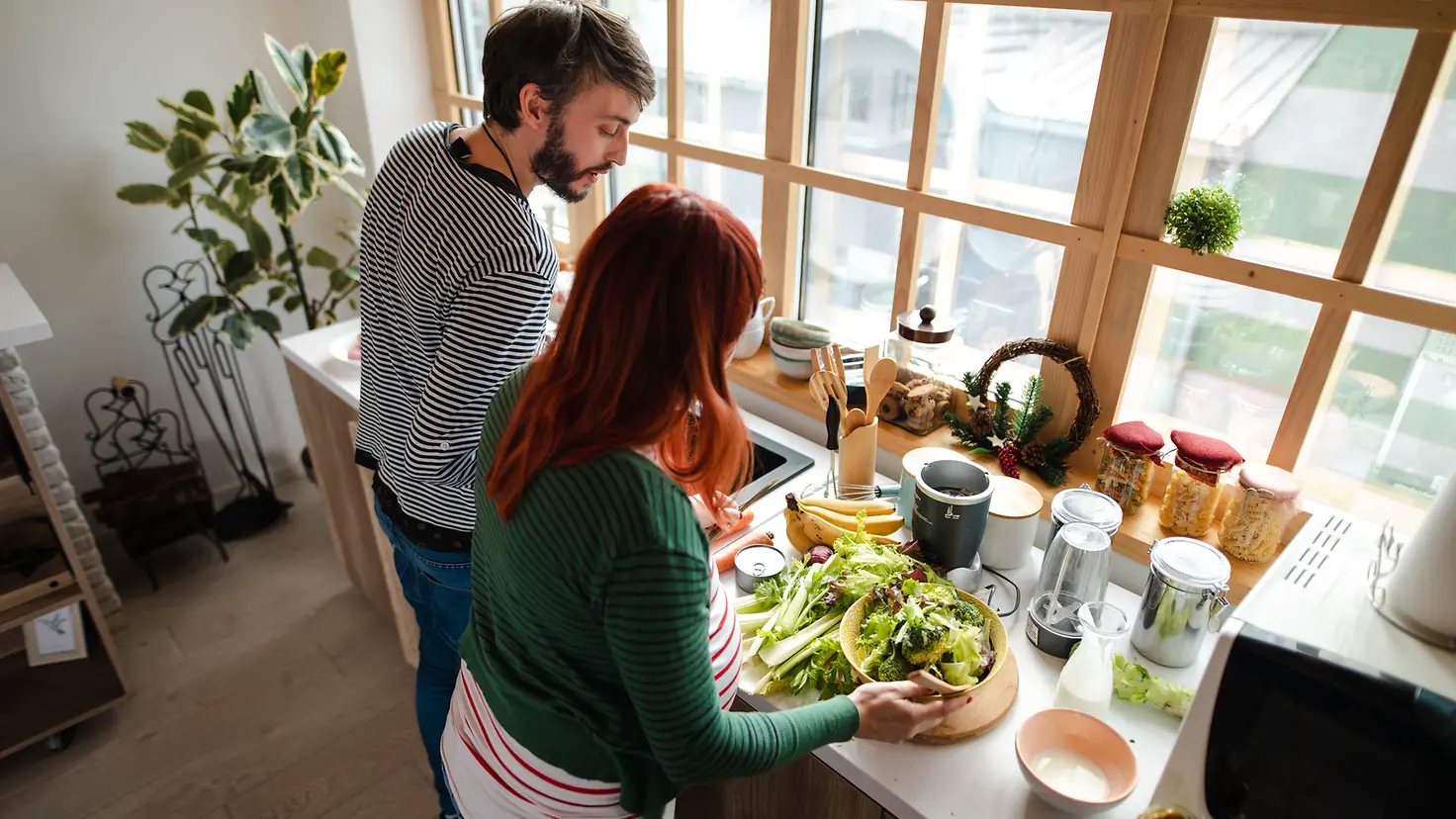 Schwangere Frau um die 29. Schwangerschaftswoche und Mann stehen in der Küche an der Arbeitsplatte und bereiten einen Salat zu