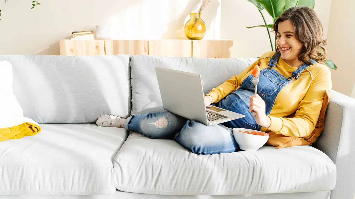 Schwangere Frau um die 29. Schwangerschaftswoche sitzt mit Laptop und einer Schüssel Obst auf der Couch