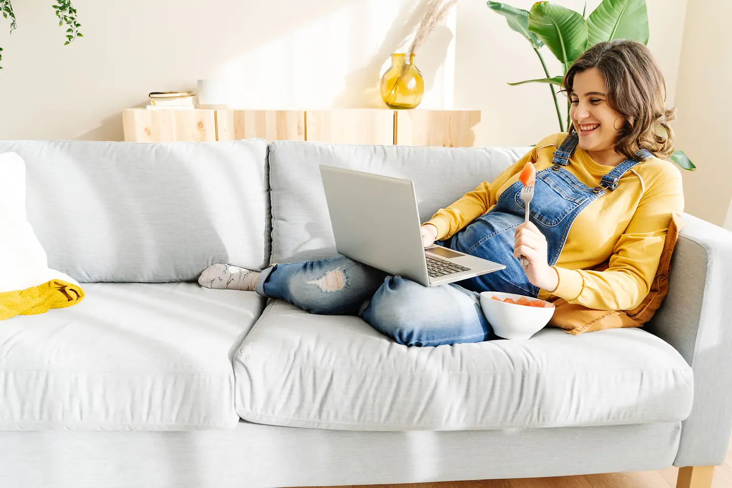 Schwangere Frau um die 29. Schwangerschaftswoche sitzt mit Laptop und einer Schüssel Obst auf der Couch