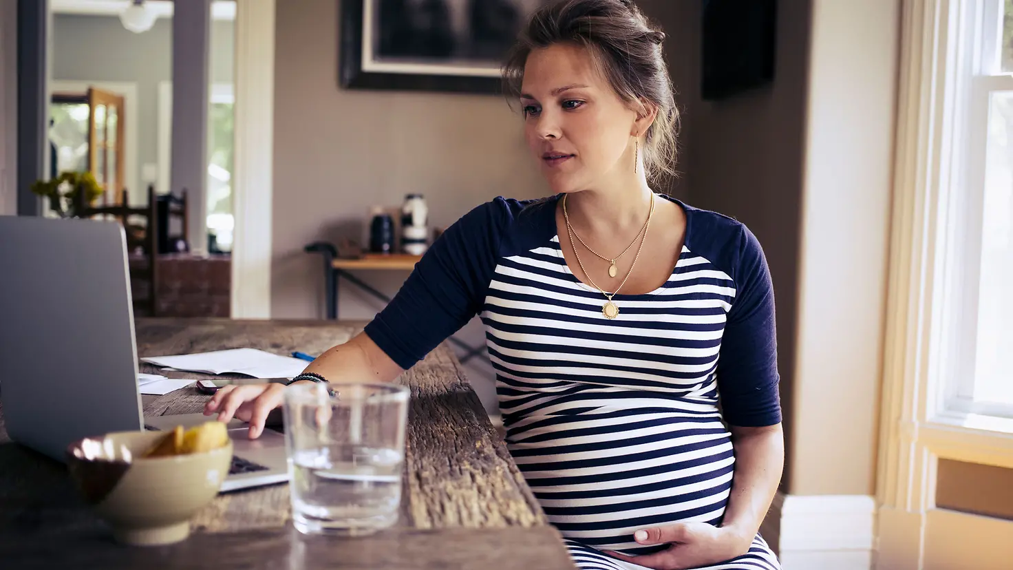 Schwangere Frau in der etwa 28. Schwangerschaftswoche sitzt an einem Esstisch, hält Ihren Bauch und schaut in einen Laptop