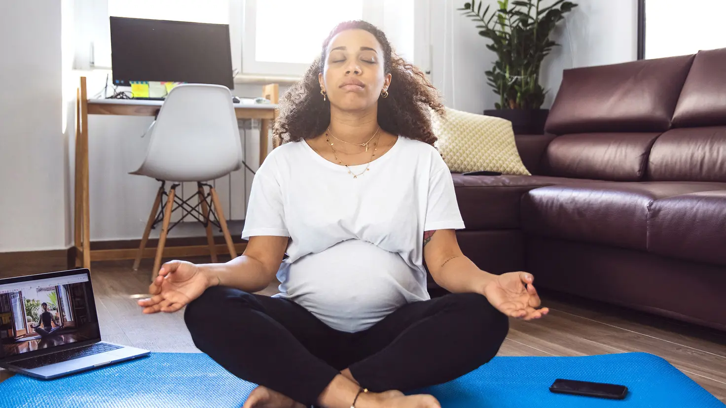Schwangere Frau in der etwa 28. Schwangerschaftswoche meditiert im Schneidersitz auf einer Jogamatte