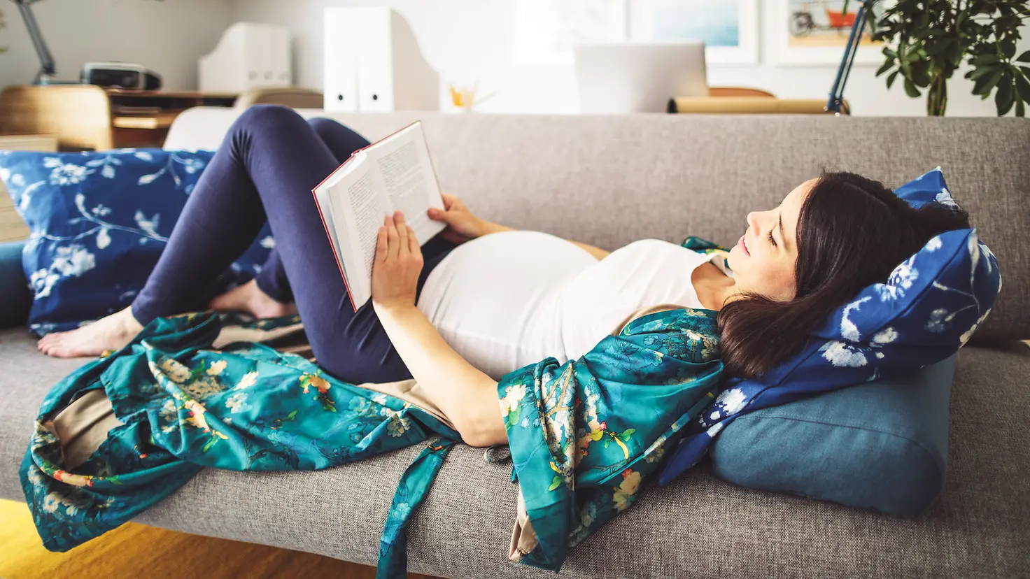 Eine junge schwangere Frau um die 31. Schwangerschaftswoche liegt auf dem Sofa und liest ein Buch