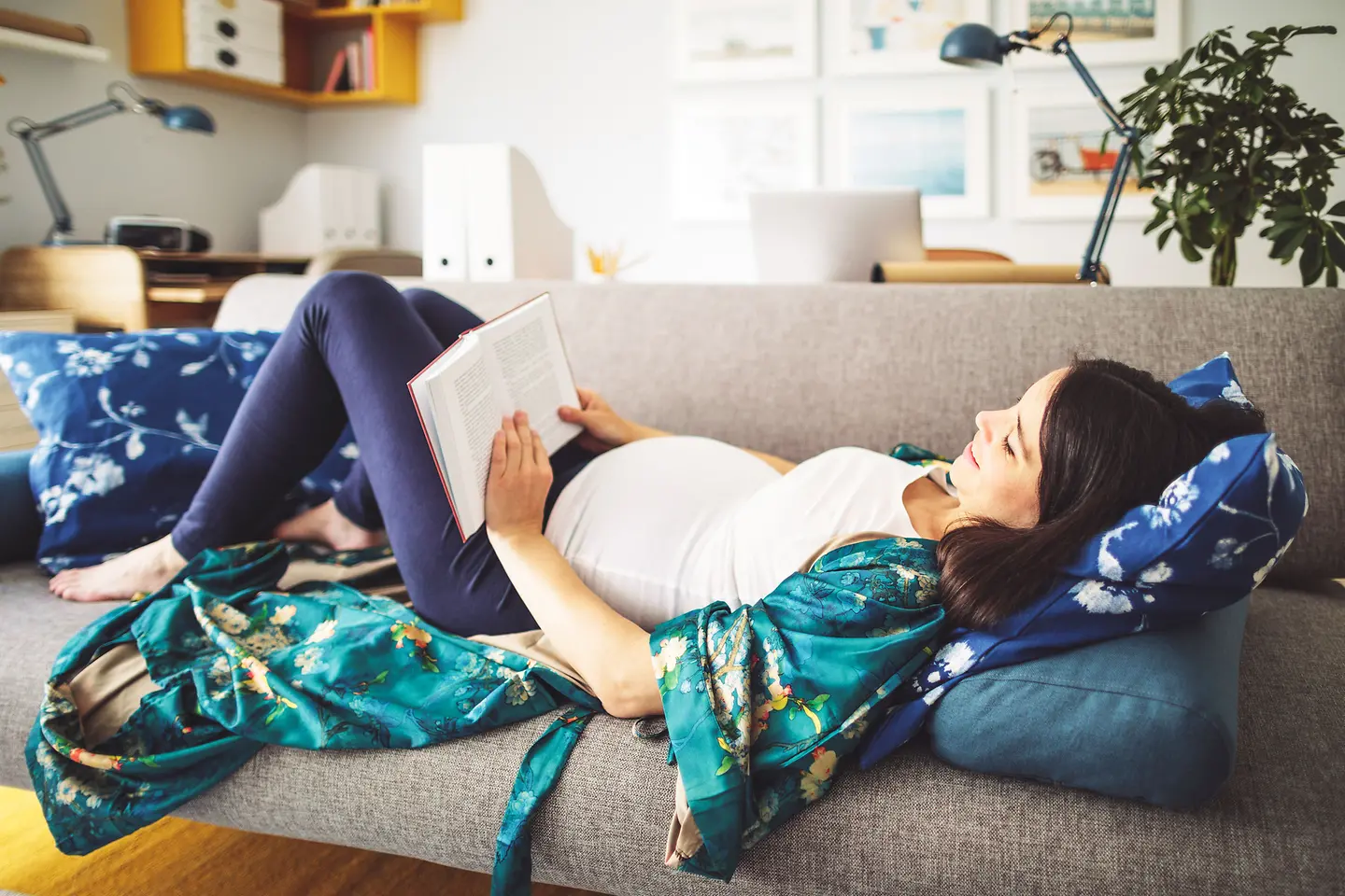 Eine junge schwangere Frau um die 31. Schwangerschaftswoche liegt auf dem Sofa und liest ein Buch