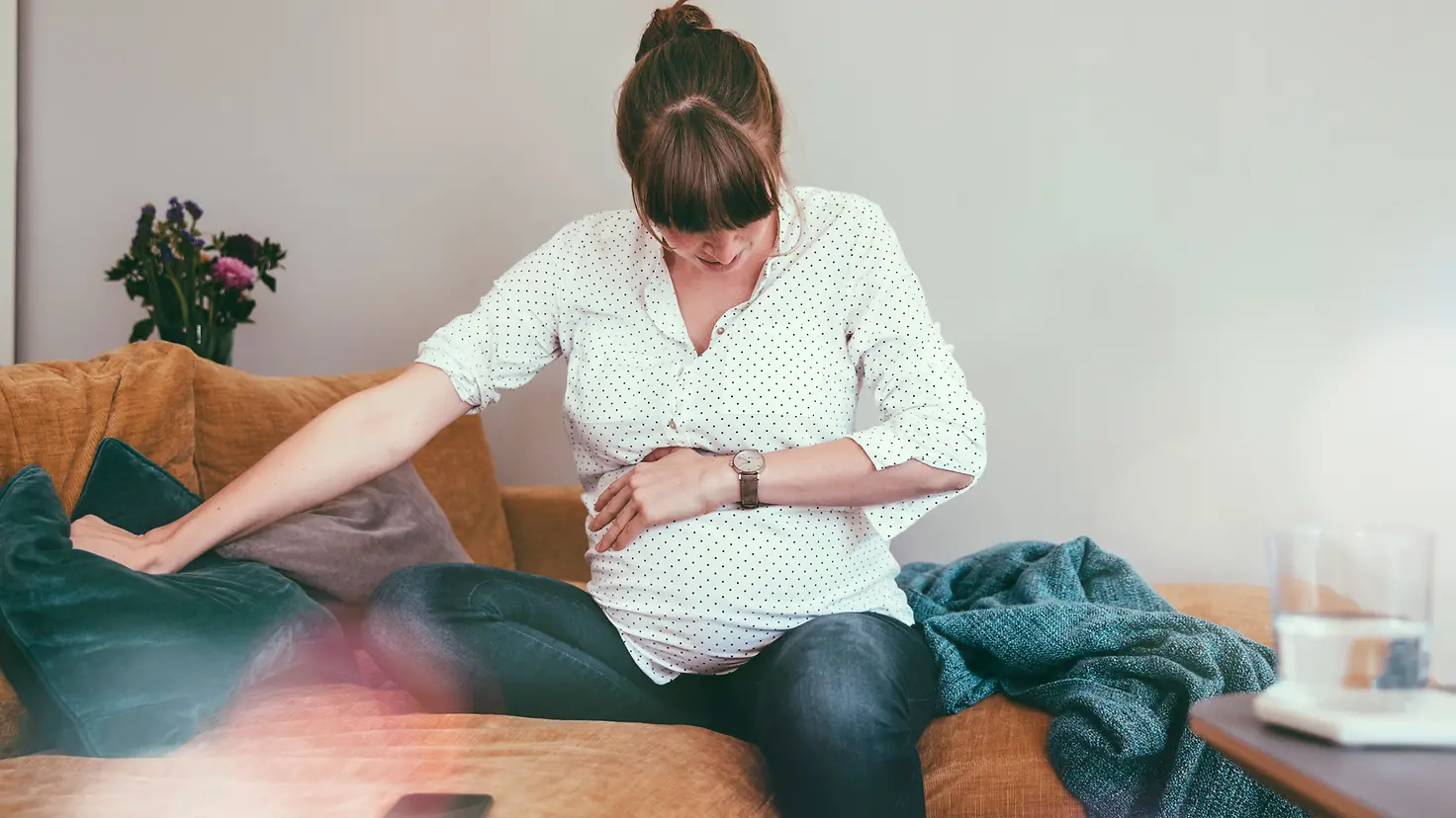 Werdende Mutter um die 32. Schwangerschaftswoche sitzt zu Hause auf der Couch, hält sich den Bauch und schaut auf ihre Armbanduhr