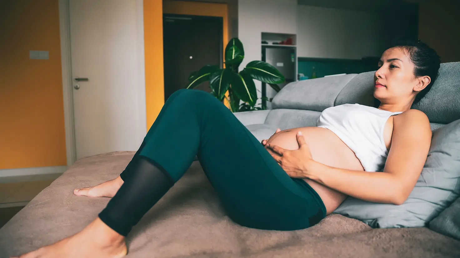 Eine schwangere Frau um die 37. Schwangerschaftswoche liegt auf einem Bett und hält sich den unteren Babybauch