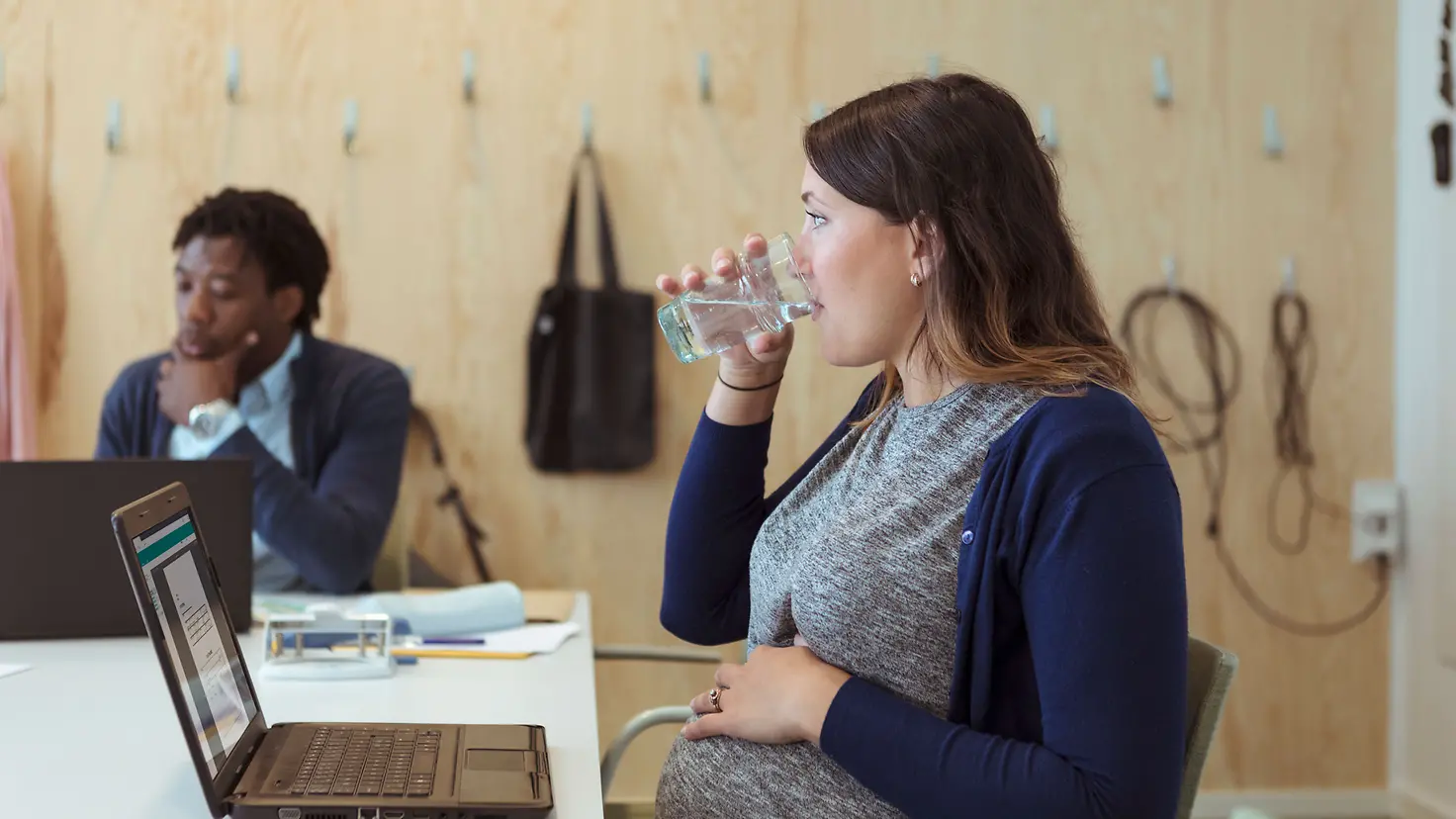 Schwangere junge Frau um die 36. Schwangerschaftswoche sitzt mit Notebook an einem Konferenztisch und trinkt ein Glas Wasser.
