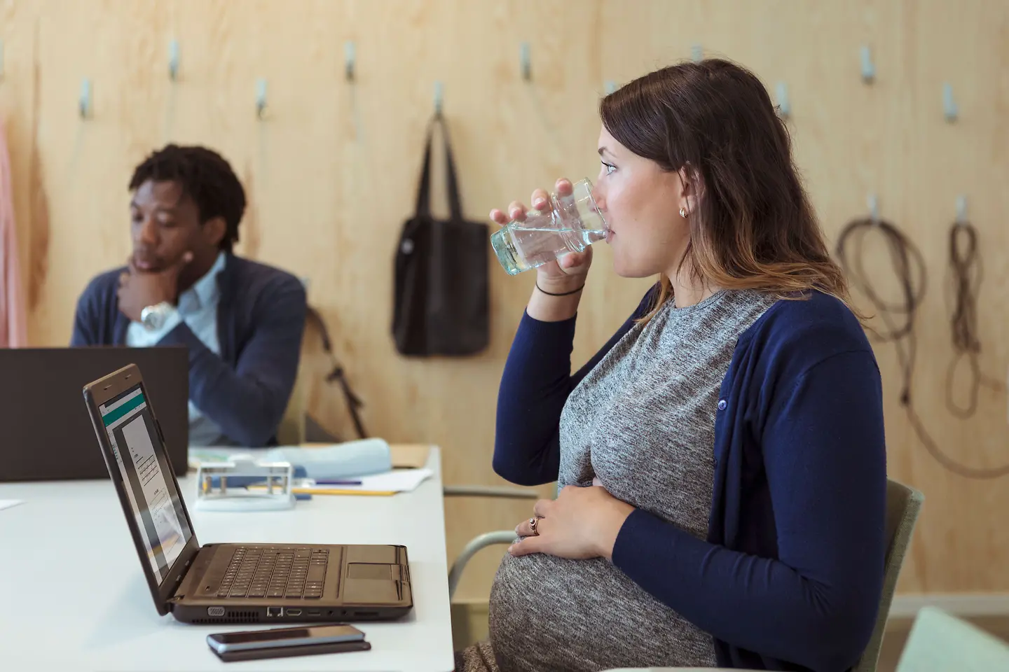 Schwangere junge Frau um die 36. Schwangerschaftswoche sitzt mit Notebook an einem Konferenztisch und trinkt ein Glas Wasser.