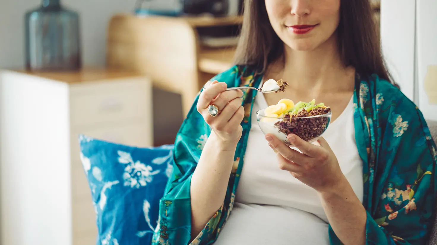 Eine schwangere Frau um die 39. Schwangerschaftswoche isst einen Becher Joghurt mit Früchten