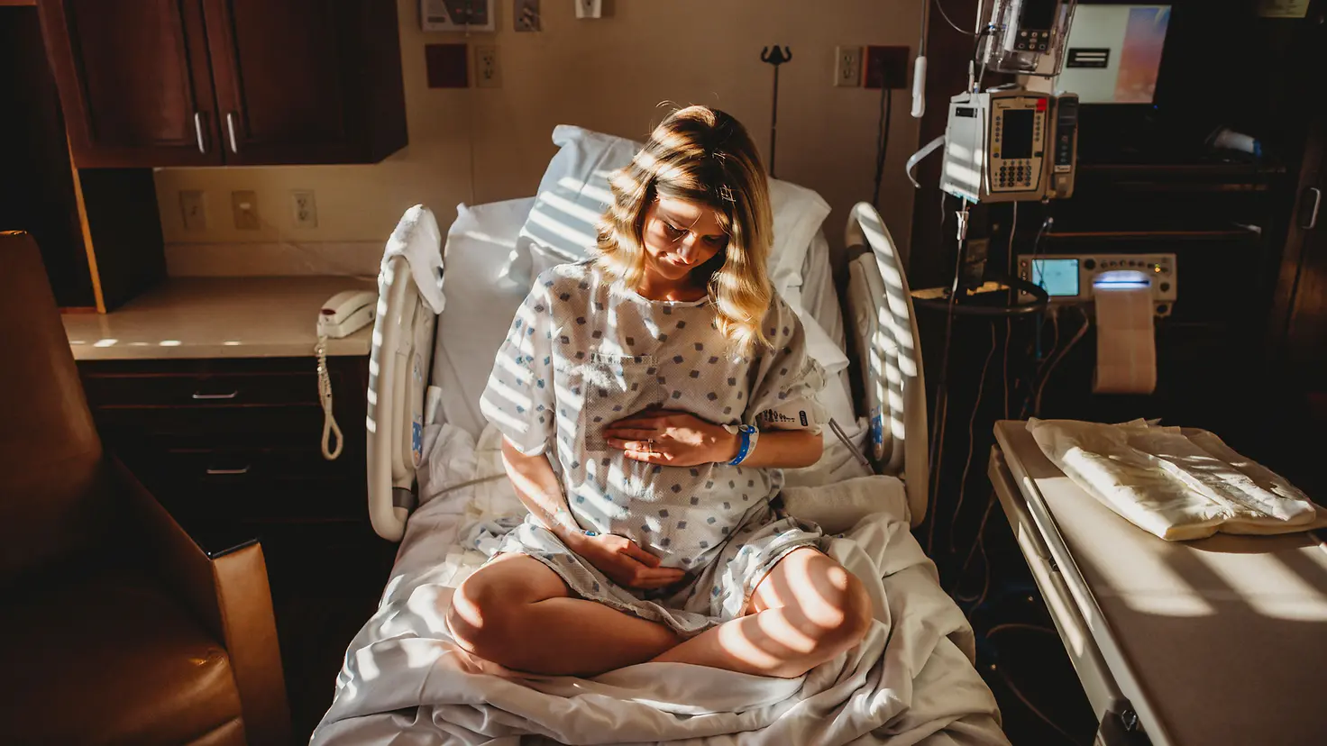 Schwangere Frau um die 40. Schwangerschaftswoche sitzt im Krankenhaus auf dem Bett und berührt ihren Bauch