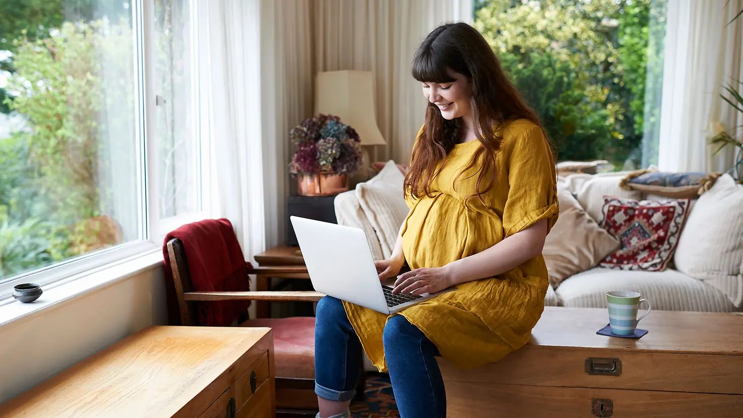 Schwangere Frau sitzt mit Laptop auf Kommode liest den Schwangerschaftsnewsletter