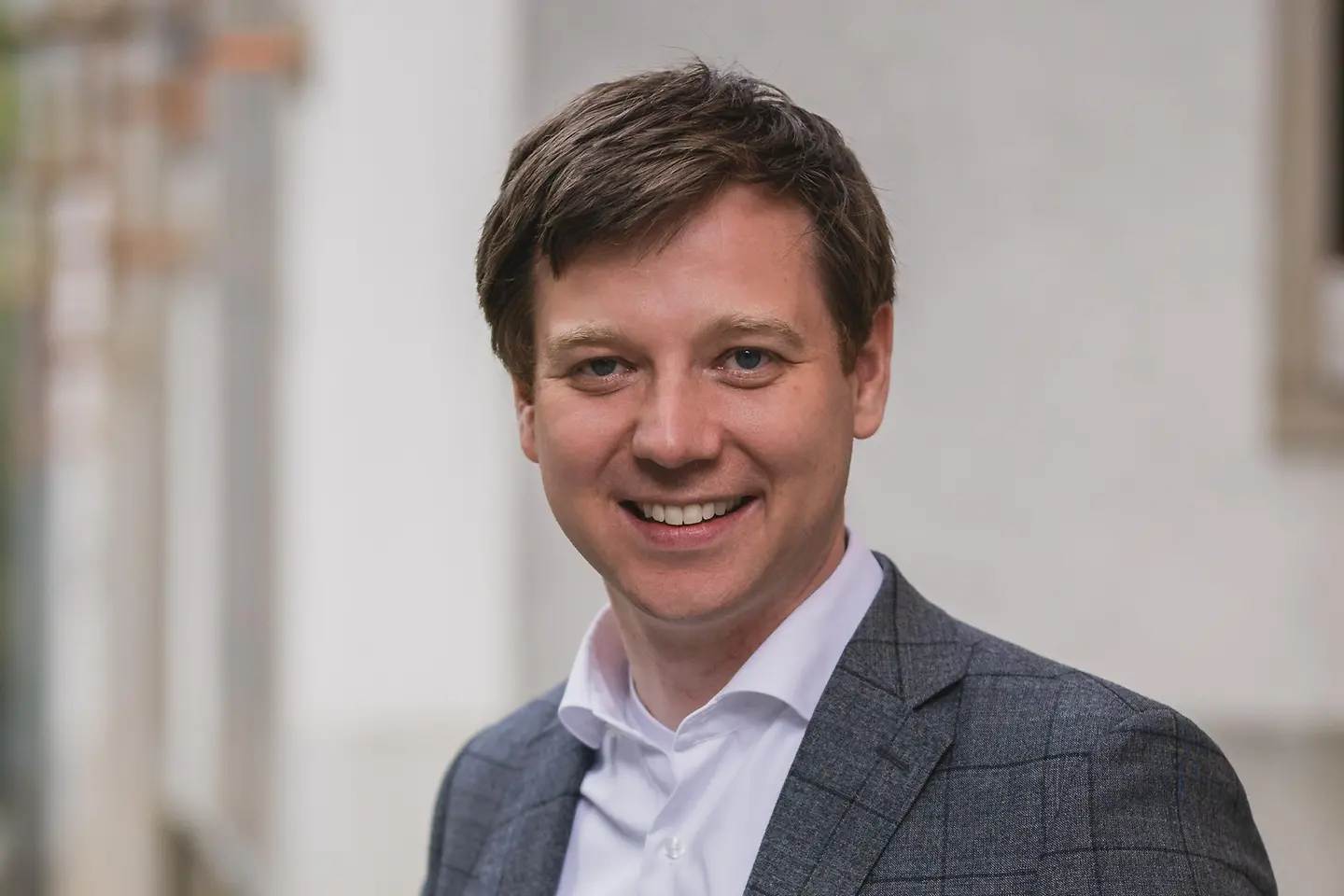 ein junger Mann mit braunem Haar, hellem Hemd und dunklem Jackett lächelt freundlich in die Kamera