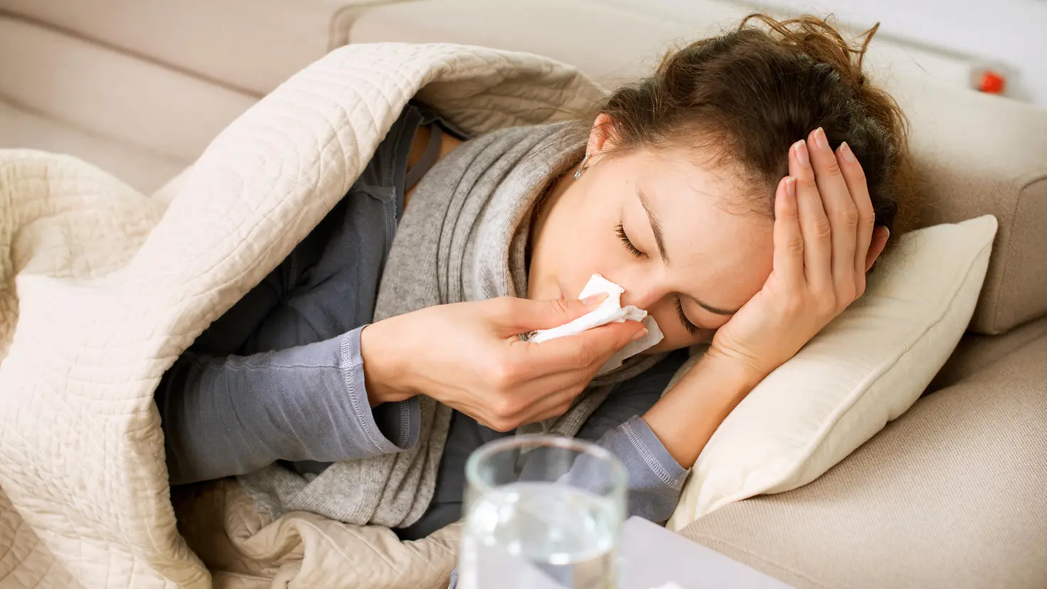 Eine Frau mit Grippe liegt auf dem Sofa und schnäuzt ihre Nase.