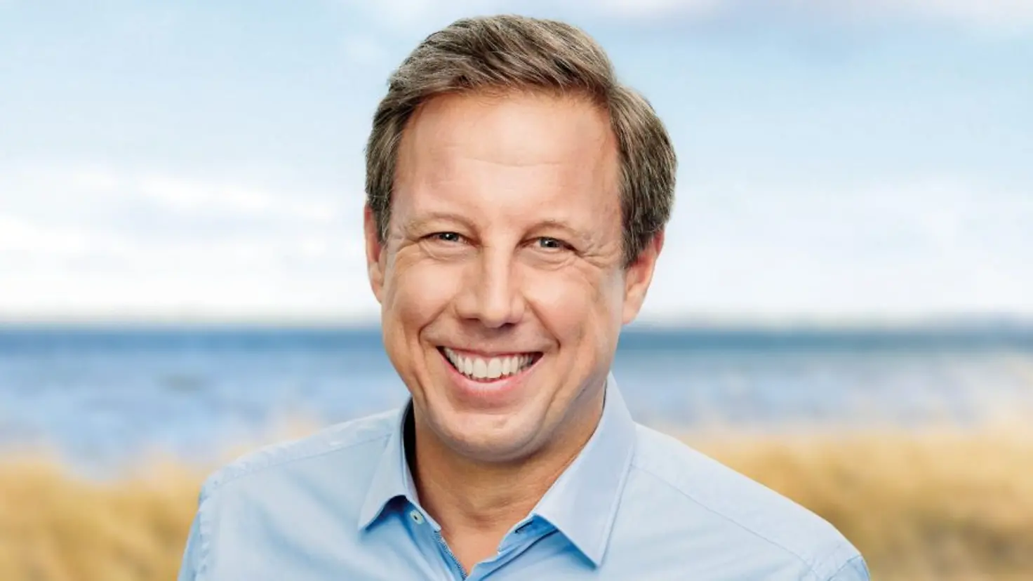 ein Mann mit hellblauem Hemd steht in den Dünen am Meer und lächelt in die Kamera