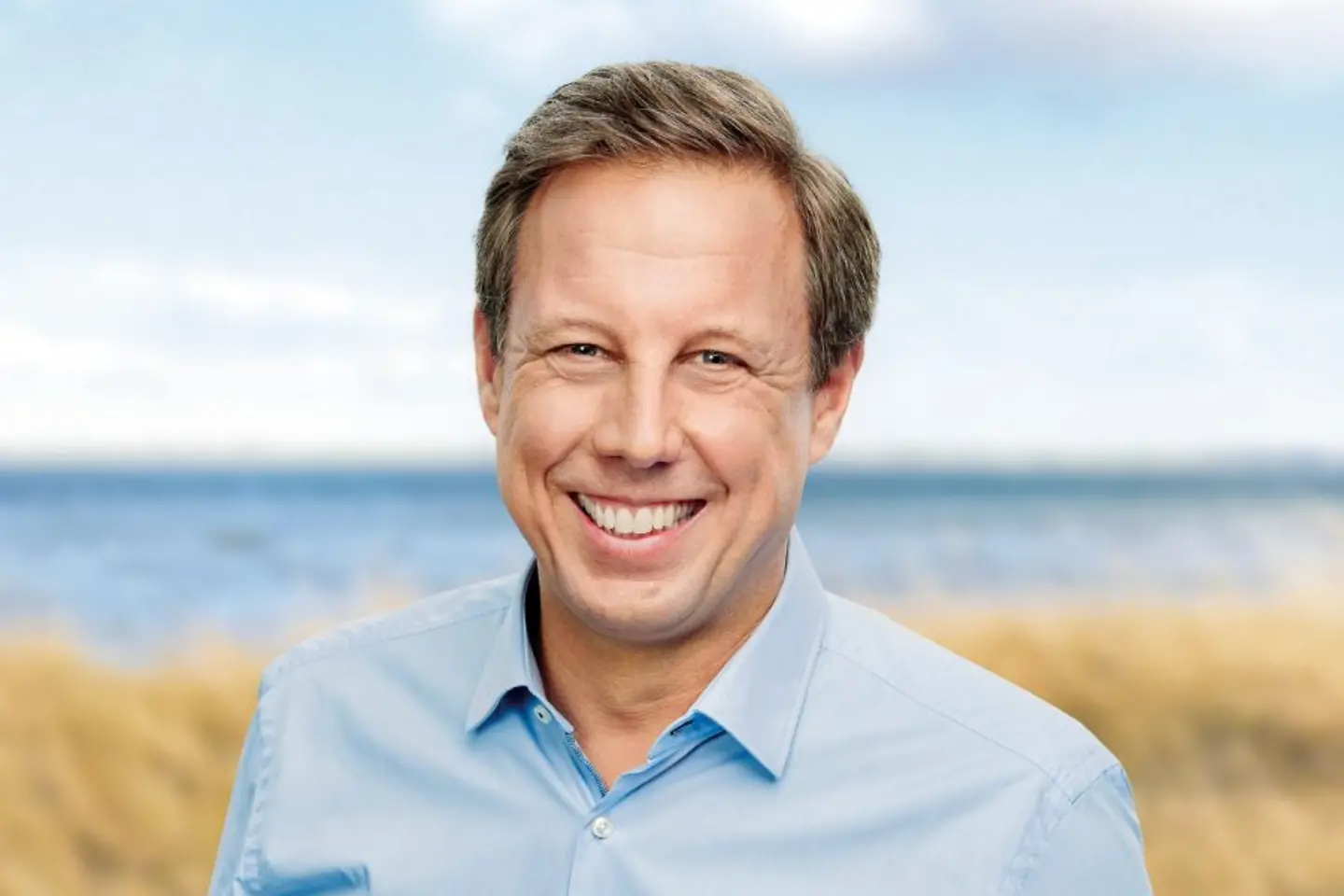 ein Mann mit hellblauem Hemd steht in den Dünen am Meer und lächelt in die Kamera