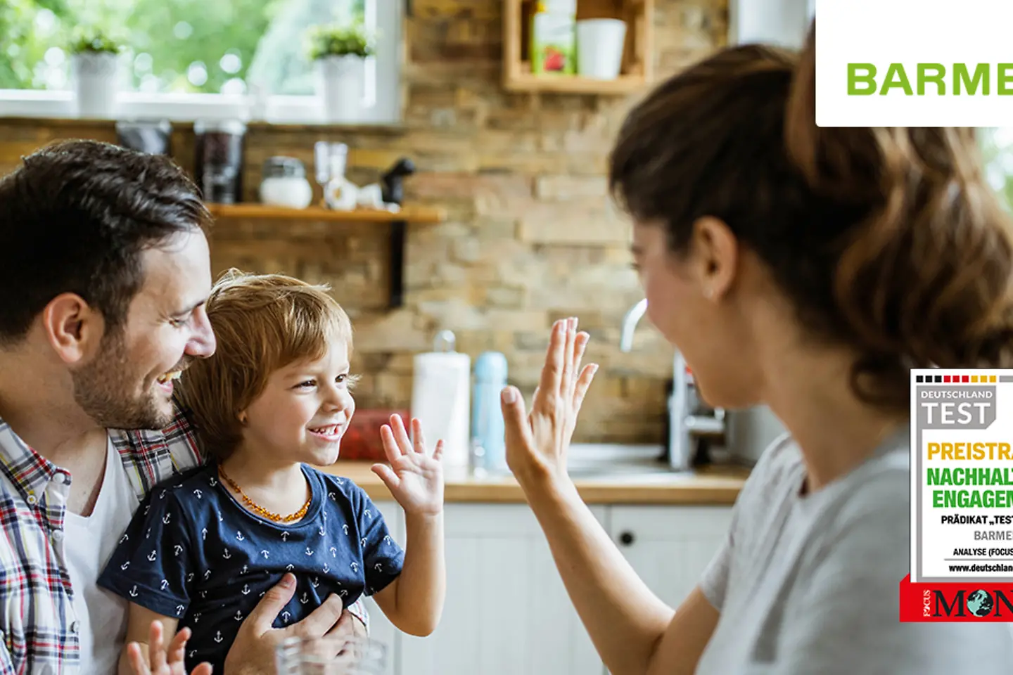 Eine junge Familie sitzt in der Küche zusammen. Mutter und Kleinkind klatschen sich ab. An der Seite sieht das Logo der Auszeichnung.