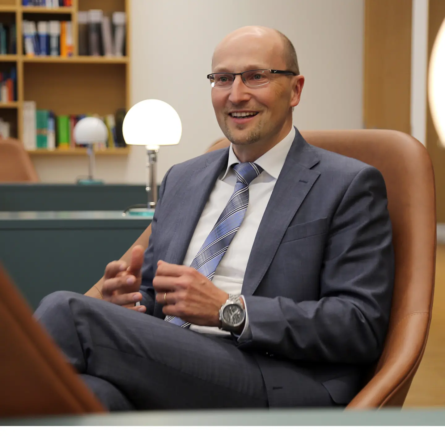 Ein Mann mit Brille, Glatze, Anzug und Krawatte sitzt in einem Sessel und spricht zu einer anderen Person.