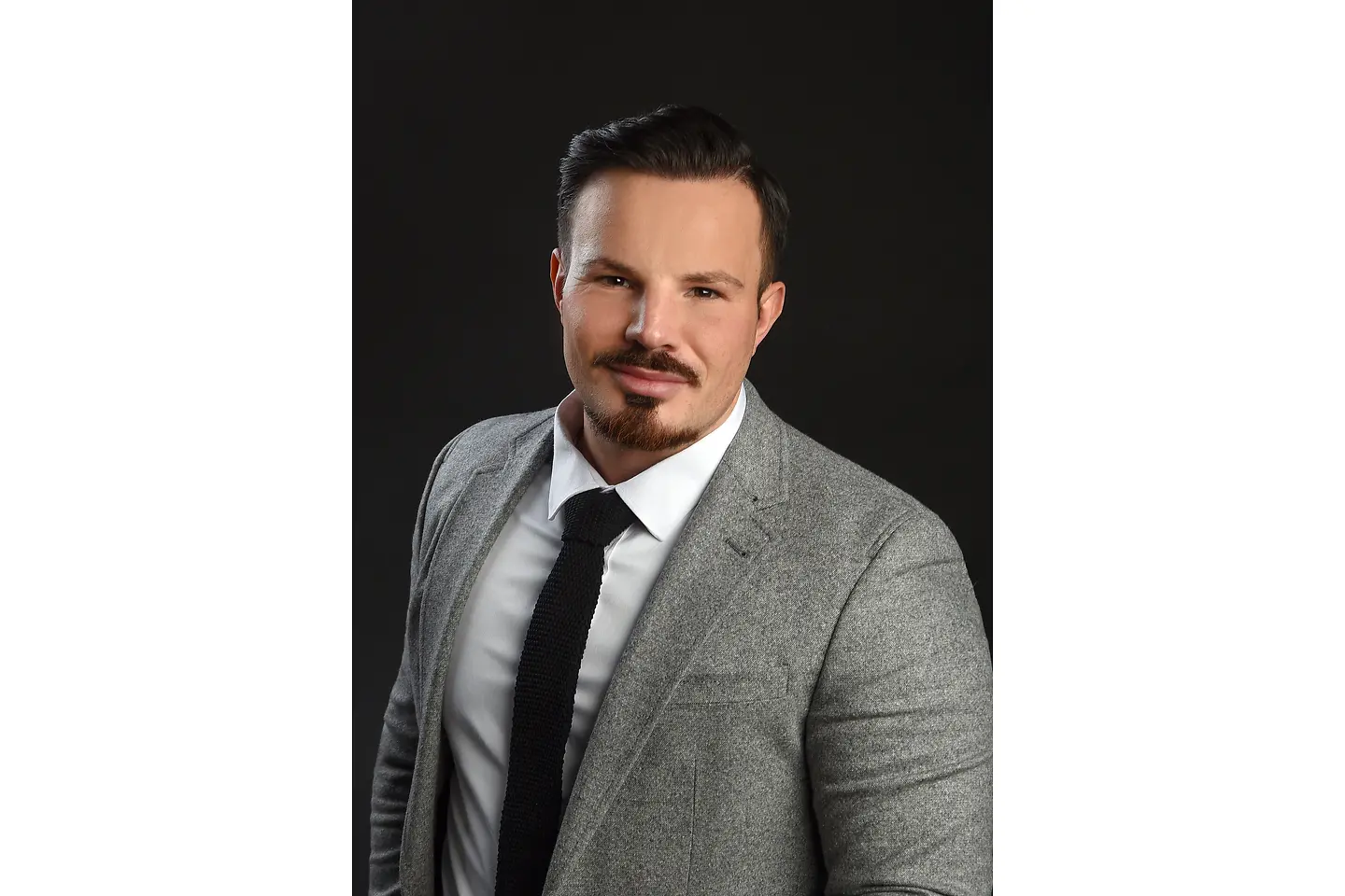 Ein junger Mann mit dunklen Haaren, grauem Sakko, Krawatte und Hemd lächelt in die Kamera