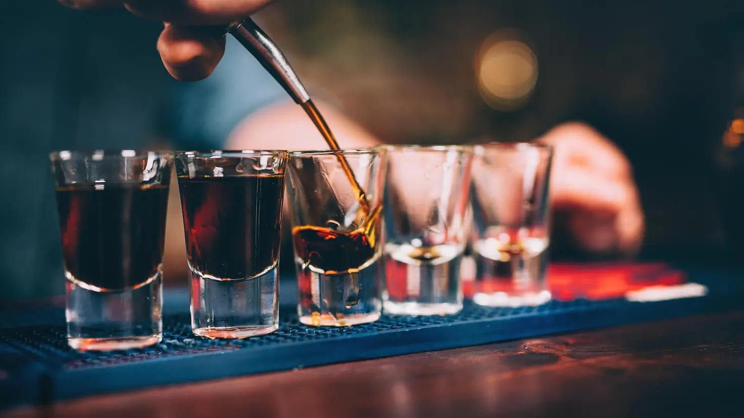 Das Bild zeigt eine Bar, auf der fünf Gläser stehen, in die ein Barkeeper Schnaps eingießt.