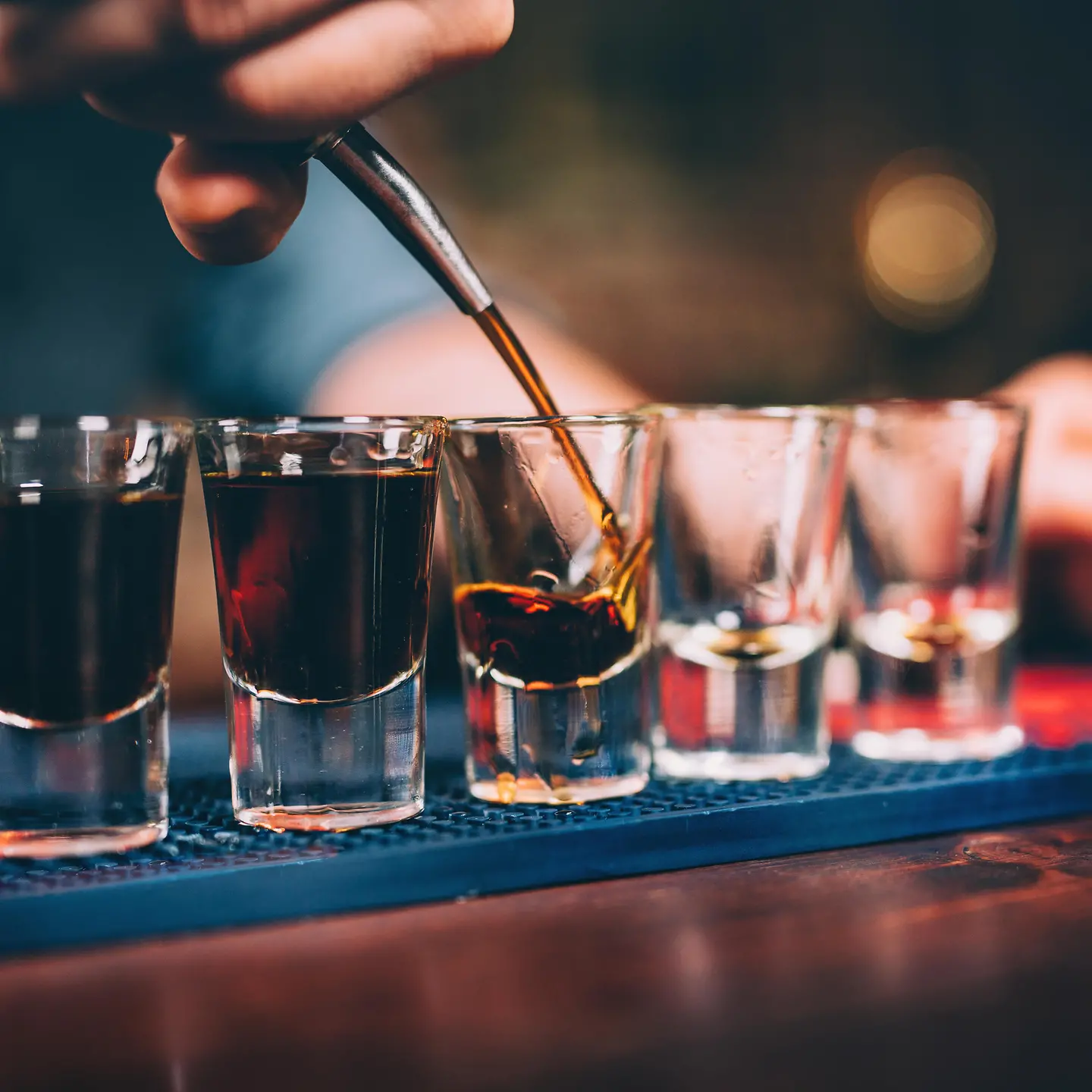 Das Bild zeigt eine Bar, auf der fünf Gläser stehen, in die ein Barkeeper Schnaps eingießt.