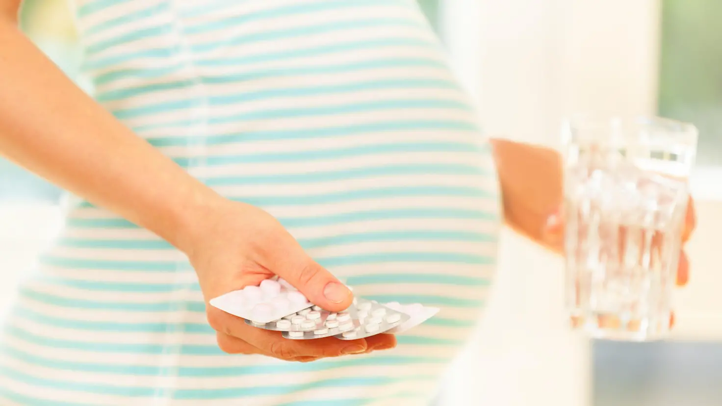 Eine schwangere Frau hält mehrere Tablettenblister in der einen Hand, in der anderen Hand ein Glas Wasser.