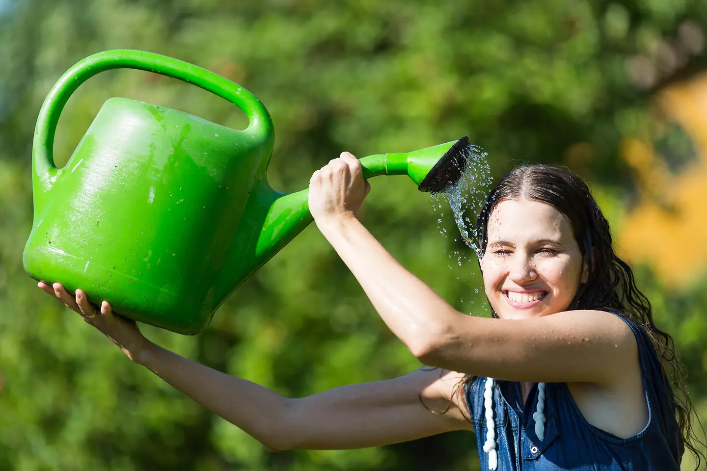 Eine lächelnde Frau gießt sich mit einer Gießkanne Wasser über den Kopf.