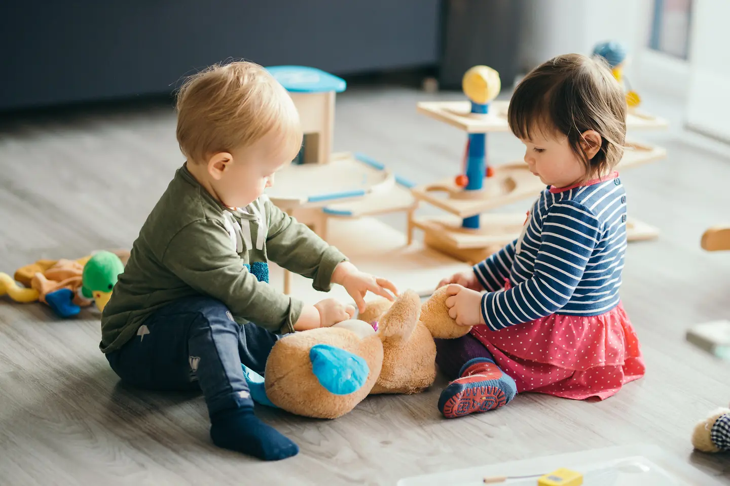 Zwei Kleinkinder sitzen auf dem Boden und spielen mit einem Teddybär.