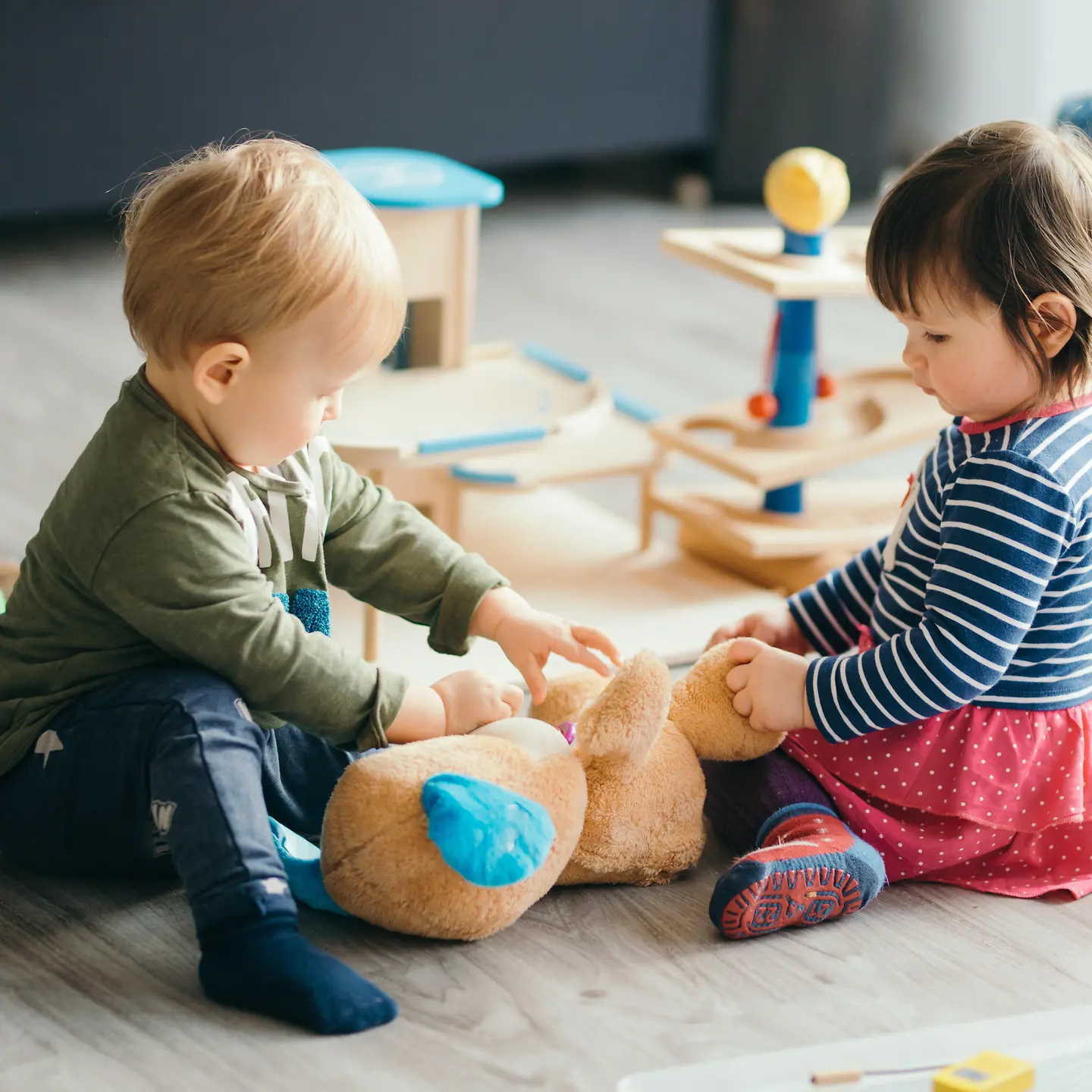 Zwei Kleinkinder sitzen auf dem Boden und spielen mit einem Teddybär.