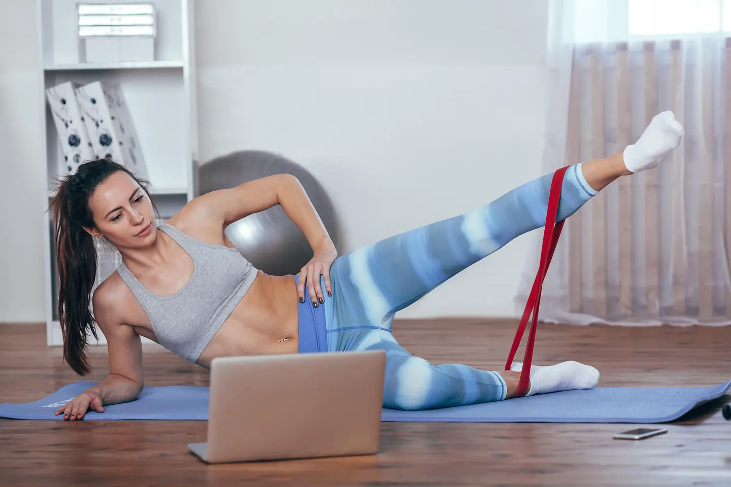Eine Frau liegt vor einem Laptop auf einer Yogamatte und macht eine Übung mit einem Fitnessband.