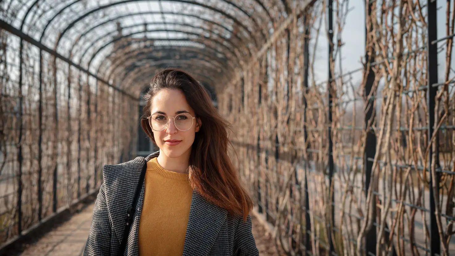 Eine junge Frau steht in einem Tunnel, der von Pflanzen umrankt wird.