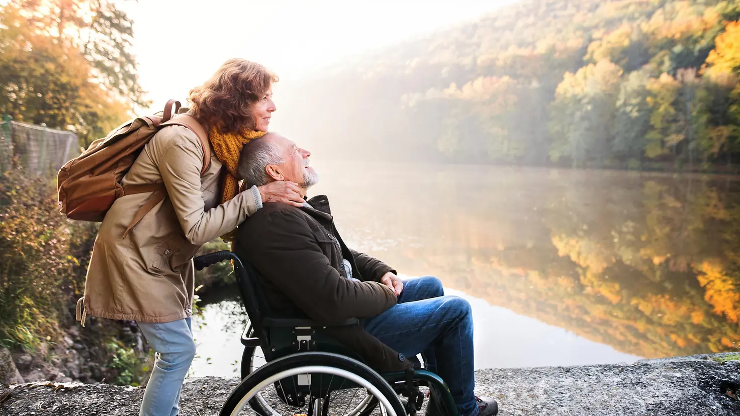 Ein älteres Paar steht an einem See, der Mann sitzt im Rollstuhl