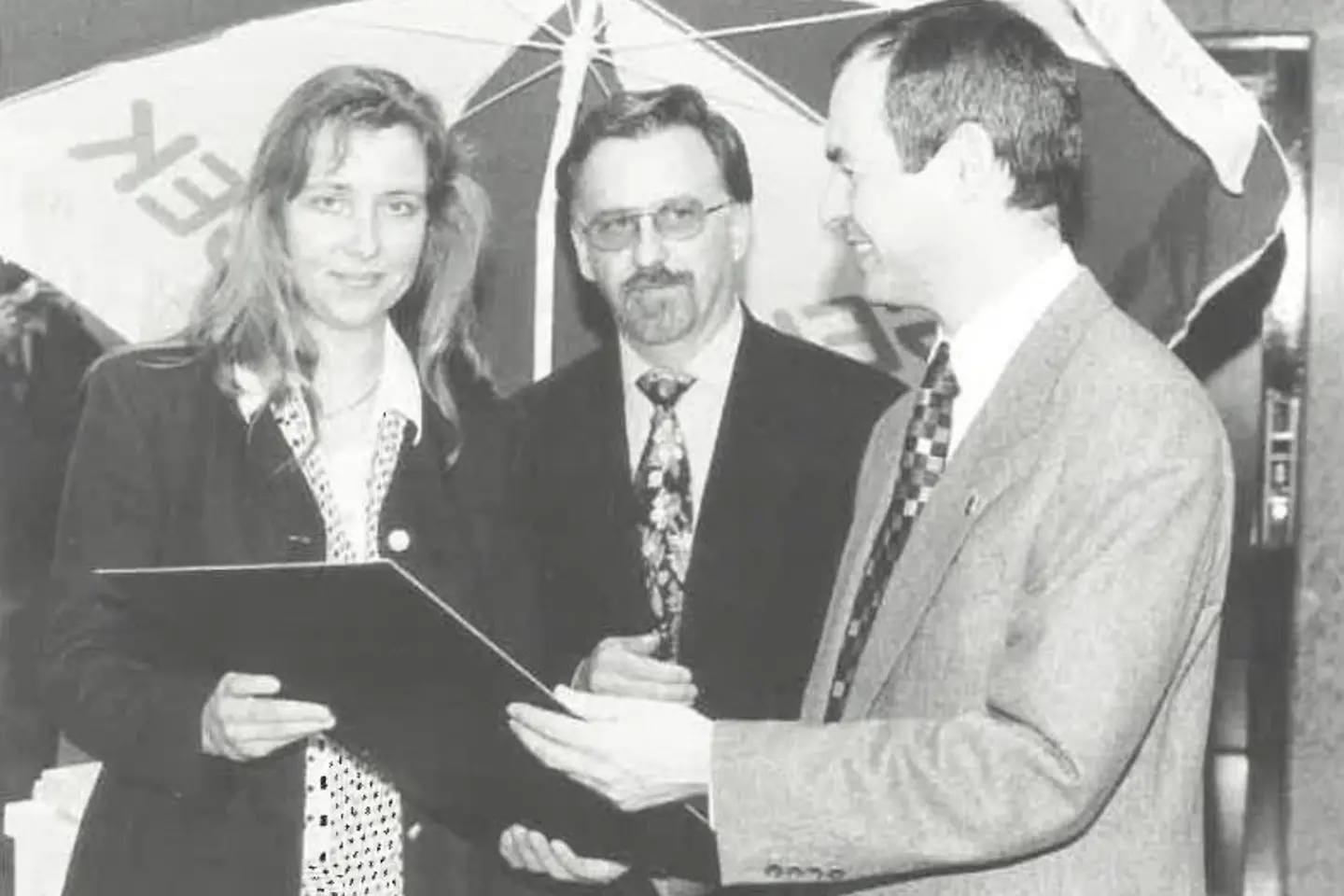 Zwei Herren im Anzug überreichen einer Frau eine Urkunde