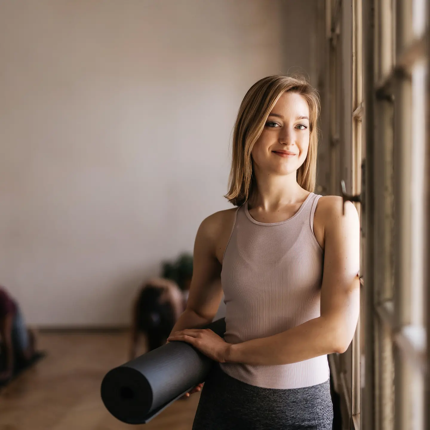 Eine junge Frau steht mit mit einer Yoganmatte in der Hand am Fenster eines Yogastudios