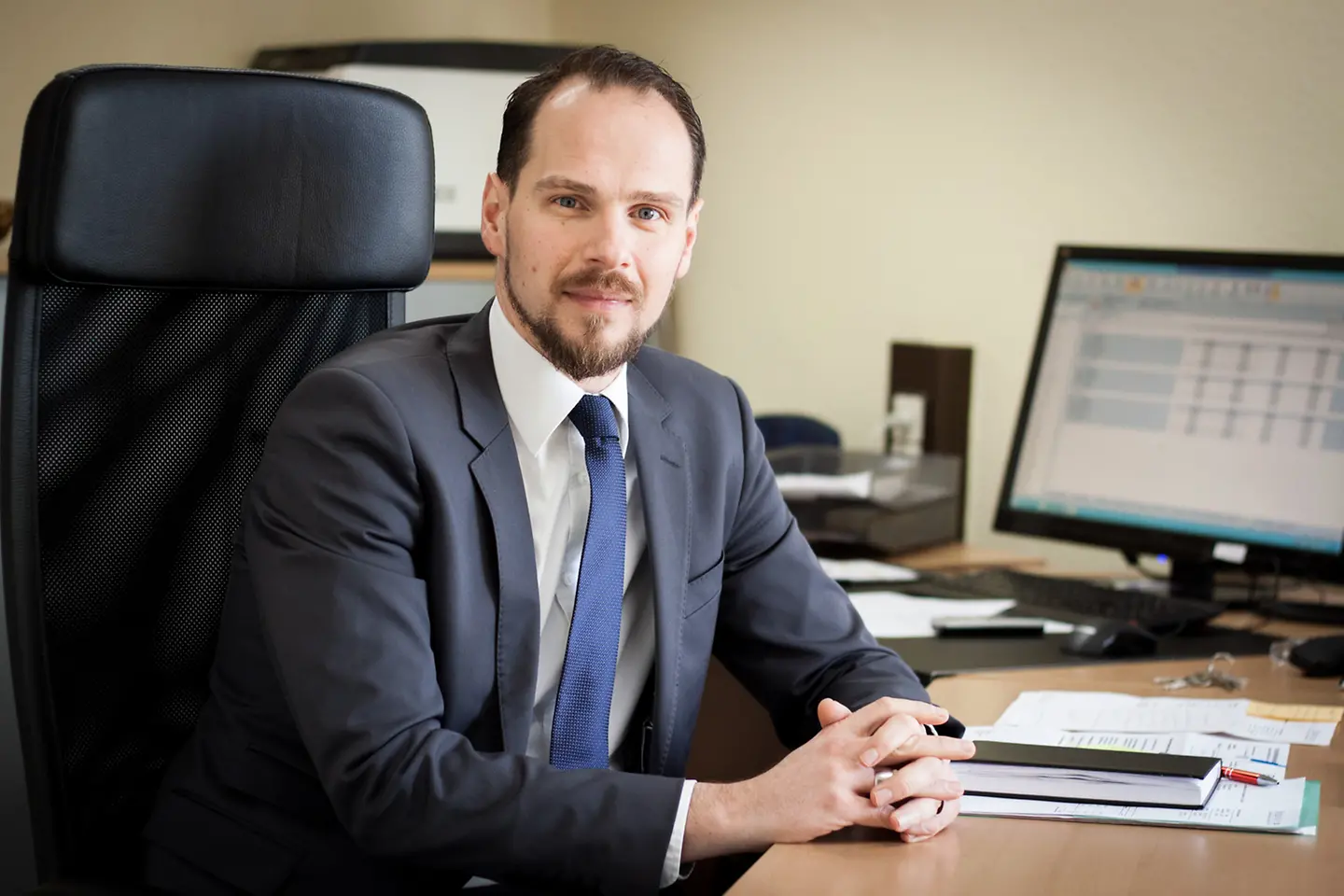 Ein junger Mann mit Bart trägt Anzug und Krawatte. Er sitzt in einem Bürostuhl und schaut in die Kamera.