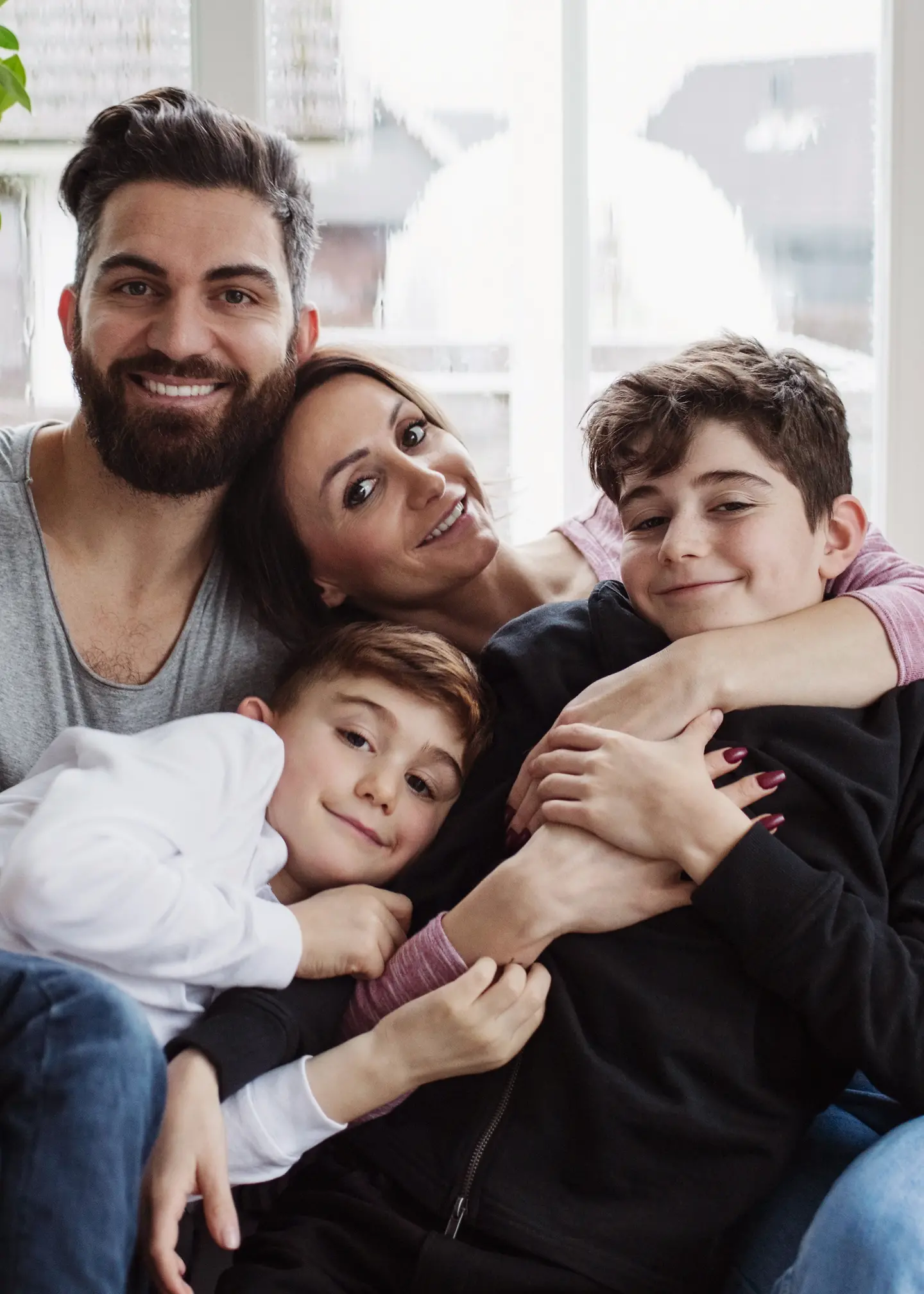 A family (two adults and two children) are sitting together on a couch, cuddling and smiling at the camera.