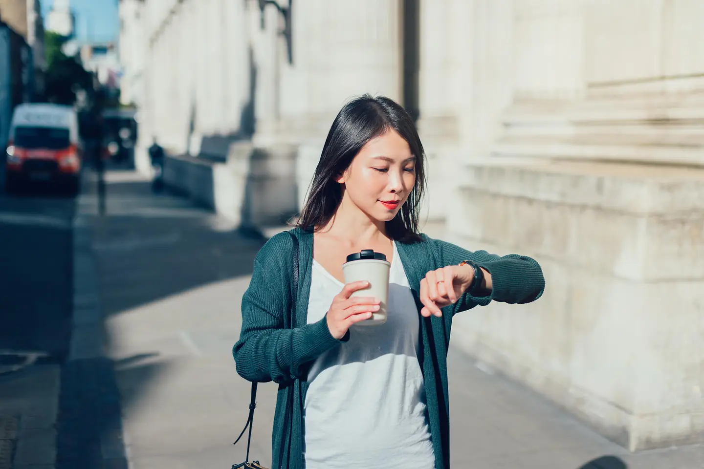 Eine junge Frau mit Kaffeebecher in der Hand schaut auf die Uhr. 