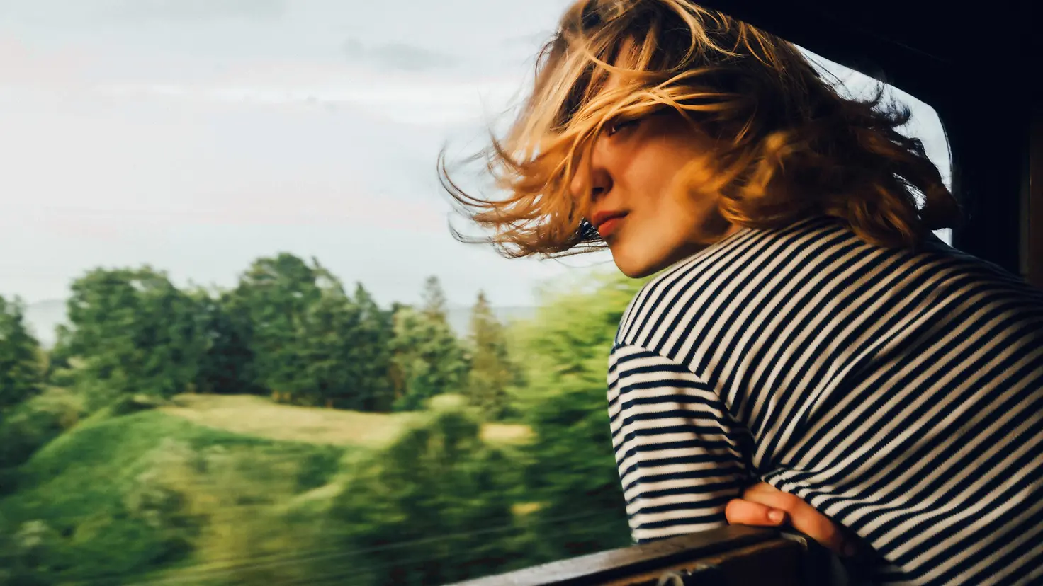 Eine junge lehnt sich aus dem geöffneten Fenster eines fahrenden Zuges und schaut in die vorbei fahrende Landschaft.