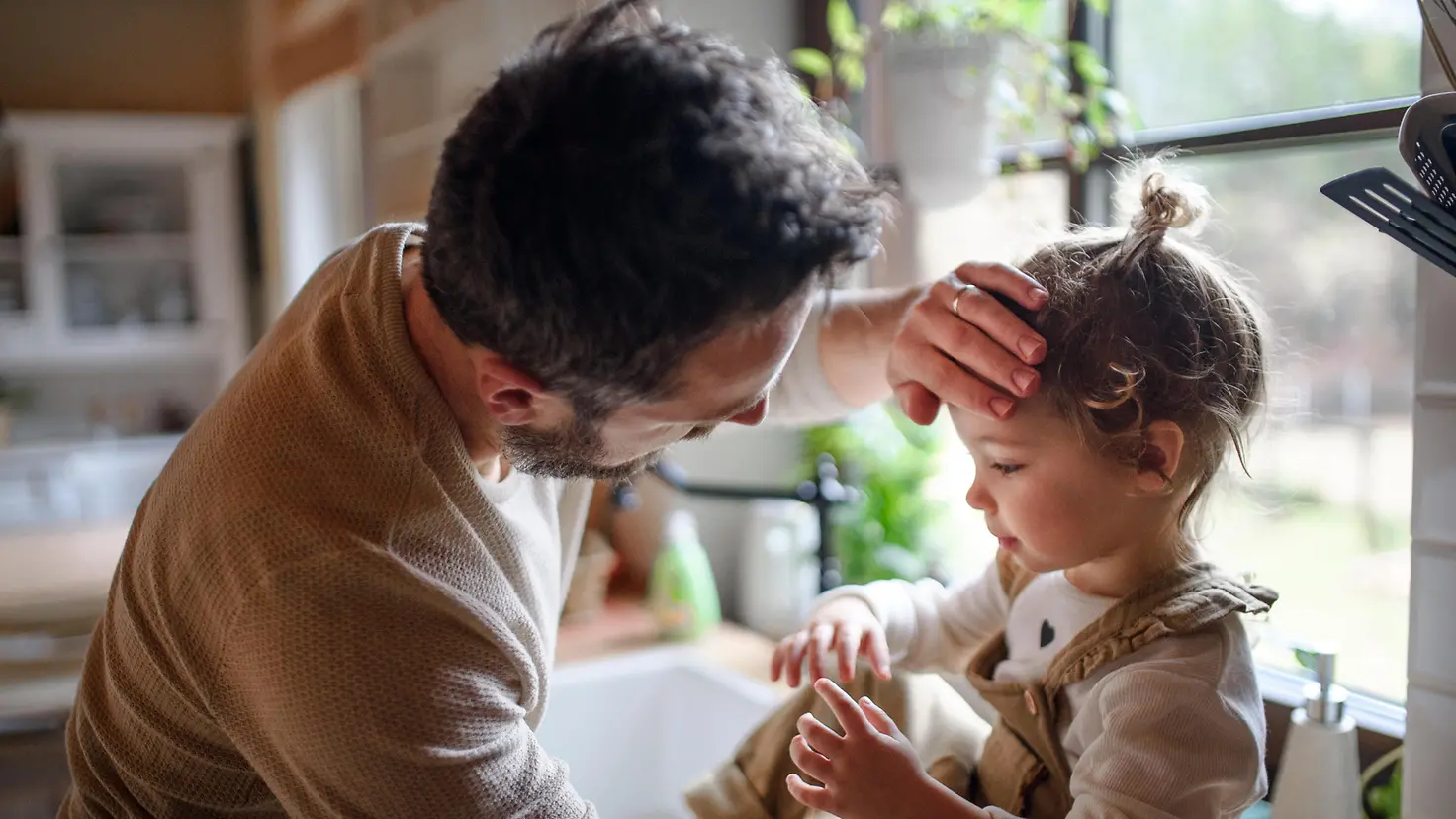 Ein Kind sitzt auf einer Küchenanrichte. Ein Mann steht vor ihm und legt seine Hand auf die Stirn des Kindes.