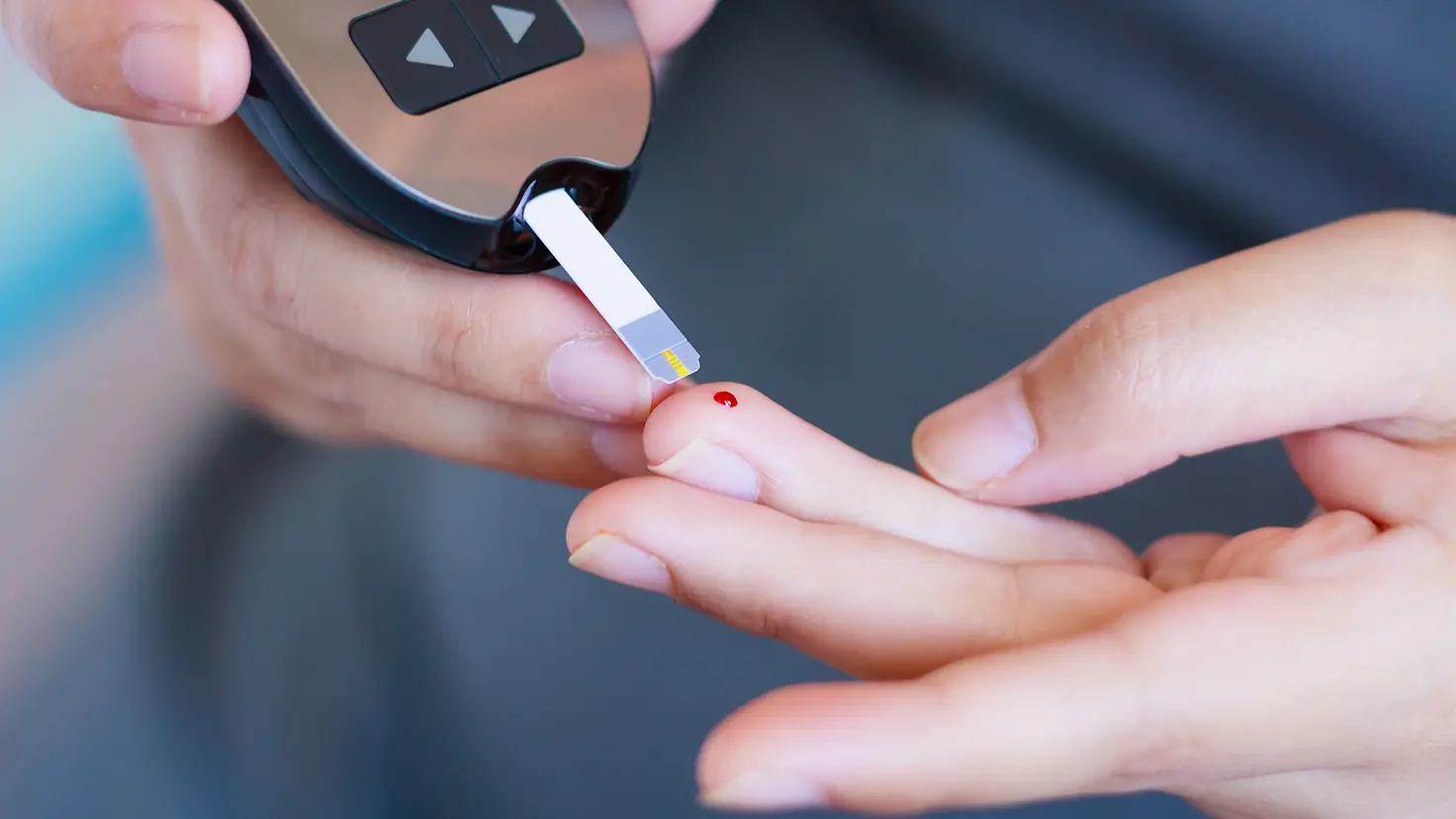 In einem Blutzucker-Messgerät steckt ein Teststreifen. Mit diesem wird ein Bluttropfen von einem Finger der anderen Hand abgenommen.