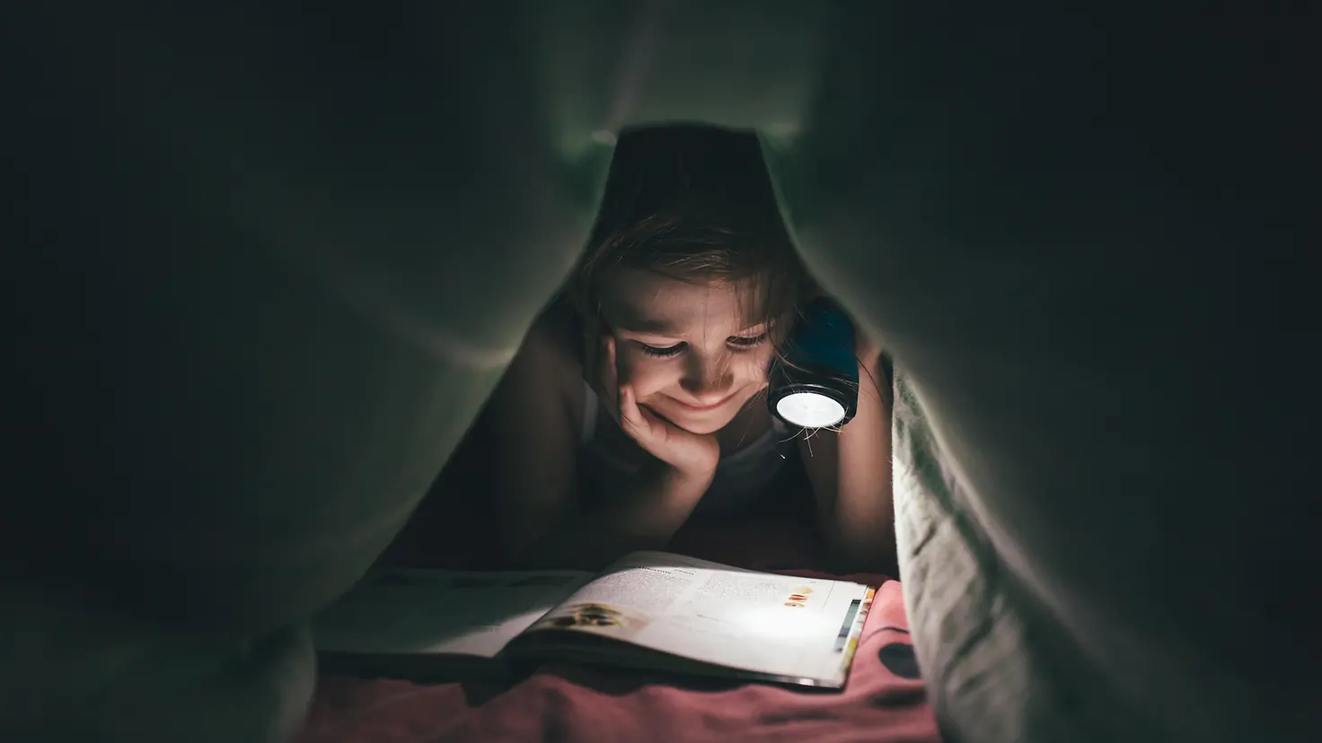 Ein Mädchen liegt im Dunkeln unter seiner Bettdecke und liest ein Buch, welches es mit einer Taschenlampe anstrahlt.chenlampe auf ein Buch