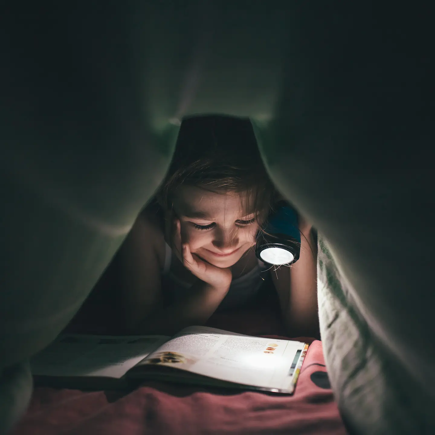 Ein Mädchen liegt im Dunkeln unter seiner Bettdecke und liest ein Buch, welches es mit einer Taschenlampe anstrahlt.chenlampe auf ein Buch